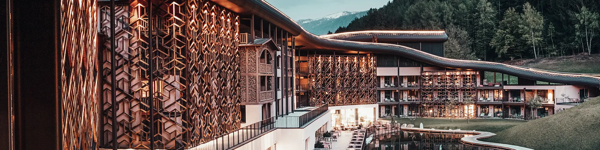 Abendaufnahme, warmes Licht beleuchtet die Holzfassade und das Aluminiumdach, unten die Terrasse mit Sitzgelegenheiten, davor der See, im Hintergrund der dunkelgrüne Wald
