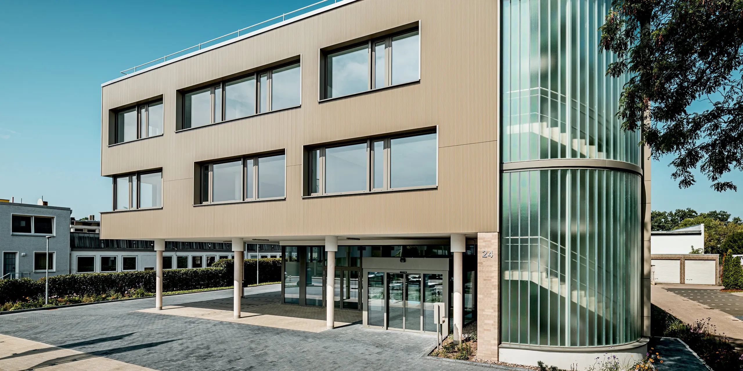 L'image présente le bâtiment Festo à Langenhagen sous un angle qui met en valeur son angle arrondi remarquable et sa façade aux profils extrudés triangle PREFA. La façade, dans une chaude teinte bronze, lui confère une allure moderne et élégante. L'angle arrondi du bâtiment est orné de surfaces vitrées verticales qui illuminent naturellement l'intérieur tout en créant une dynamique visuelle captivante. L'alliance de matériaux PREFA robustes et faciles d'entretien et de larges baies vitrées garantit à la fois esthétique et fonctionnalité. Cette conception fait de ce bâtiment un exemple remarquable d'architecture moderne qui se distingue dans son environnement.