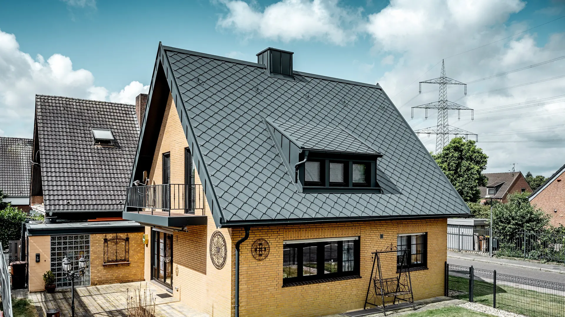 Maison individuelle en briques jaunes après la rénovation du toit avec un losange de toiture PREFA 29 × 29 en gris sombre ; toit en pente raide nouvellement recouvert avec un motif en losange, une lucarne et une cheminée intégrées, terrasse et jardin au premier plan.