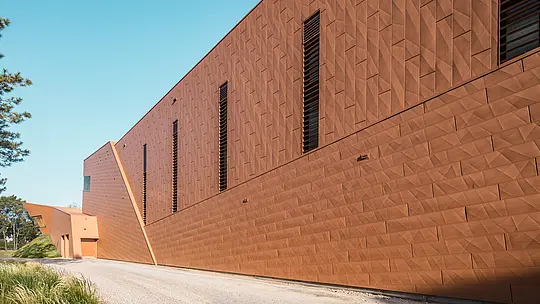 La façade dynamique et plissée de la station de pompage d'eau potable WML à Lottum se distingue par ses étroites ouvertures vitrées, judicieusement placées, qui attirent le regard. La façade en aluminium PREFA Siding.X, à l'aspect chaleureux et rouillé, crée un jeu d'ombres, de lumières et de géométrie saisissant, soulignant la pureté des lignes du bâtiment. Cette structure exceptionnelle allie fonctionnalité technique et esthétique sculpturale. Légers, durables et entièrement recyclables, les panneaux en aluminium PREFA offrent une résistance supérieure aux intempéries, une excellente stabilité dimensionnelle et une enveloppe de bâtiment durable et nécessitant peu d'entretien.