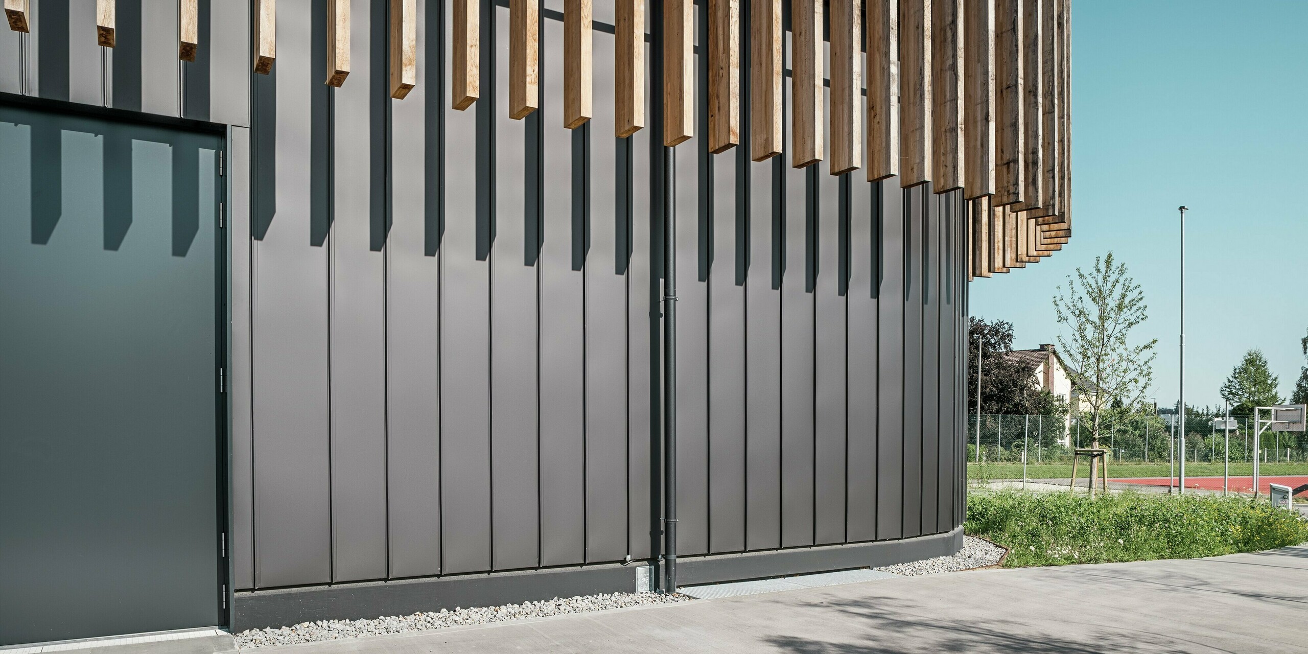Gros plan du rez-de-chaussée du Team 7 World à Ried im Innkreis : la façade PREFALZ noir-brun forme une image globale harmonieuse avec les éléments verticaux en bois. Le revêtement robuste en aluminium protège des intempéries et impressionne par sa qualité durable et sans entretien. Le jeu d'ombre et de lumière souligne les lignes claires et l'architecture moderne du complexe immobilier.