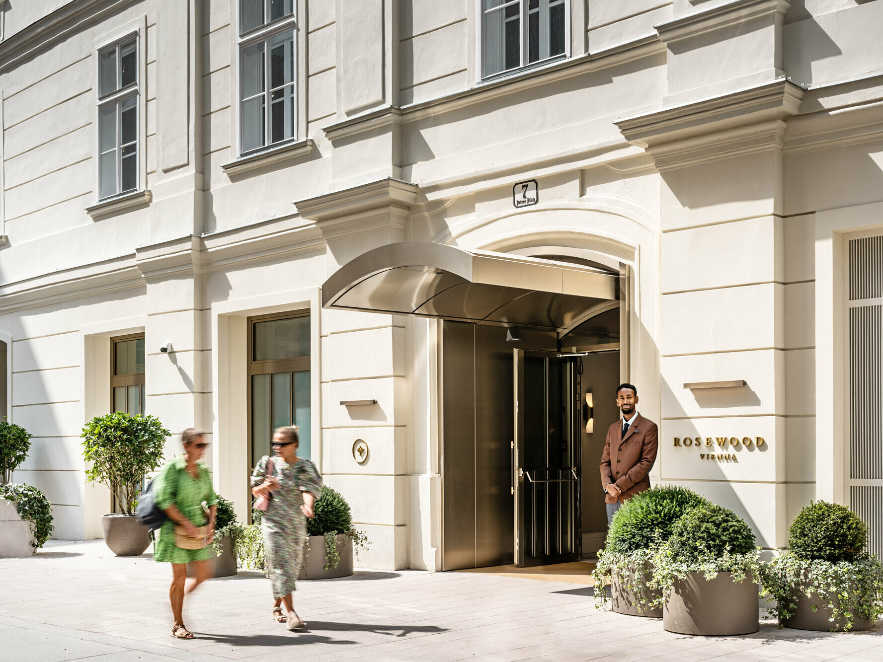 Entrée de l'hôtel Rosewood, devant laquelle on peut voir un portier, des passants et des plantes en pot de belles dimensions.