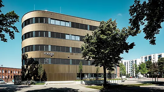 Photographie architecturale dynamique du commissariat 43 à Hambourg : le nouveau bâtiment incurvé, avec sa façade en aluminium PREFA composée de profils extrudés « triangle  » couleur bronze moyen, attire immédiatement le regard dans le paysage urbain. La surface finement texturée crée de magnifiques jeux d’ombre et de lumière entre les bandes horizontales de fenêtres et souligne les lignes fluides de la façade. L’aluminium PREFA séduit par sa haute résistance à la corrosion, sa durabilité et ses faibles besoins d’entretien ; il est idéal pour des solutions de façade durables, fonctionnelles et d’une grande précision architecturale.