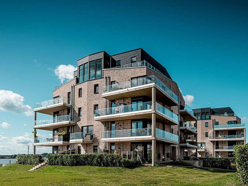 Le complexe résidentiel « Hafen zur Freiheit » (Port vers la liberté) à Schleswig présente une architecture moderne en bord de mer : des façades en briques claires côtoient des éléments Siding.X PREFA P.10 anthracite à la structure dynamique. Les étages supérieurs, particulièrement remarquables, se distinguent par leurs généreuses balustrades en verre et leurs lignes épurées qui créent un élégant contraste sous la lumière du soleil. Les façades en aluminium résistant assurent une protection durable contre les intempéries côtières et confèrent au complexe un caractère distinctif et haut de gamme. Les balcons spacieux offrent une vue imprenable sur le paysage maritime et contribuent à un confort de vie optimal.