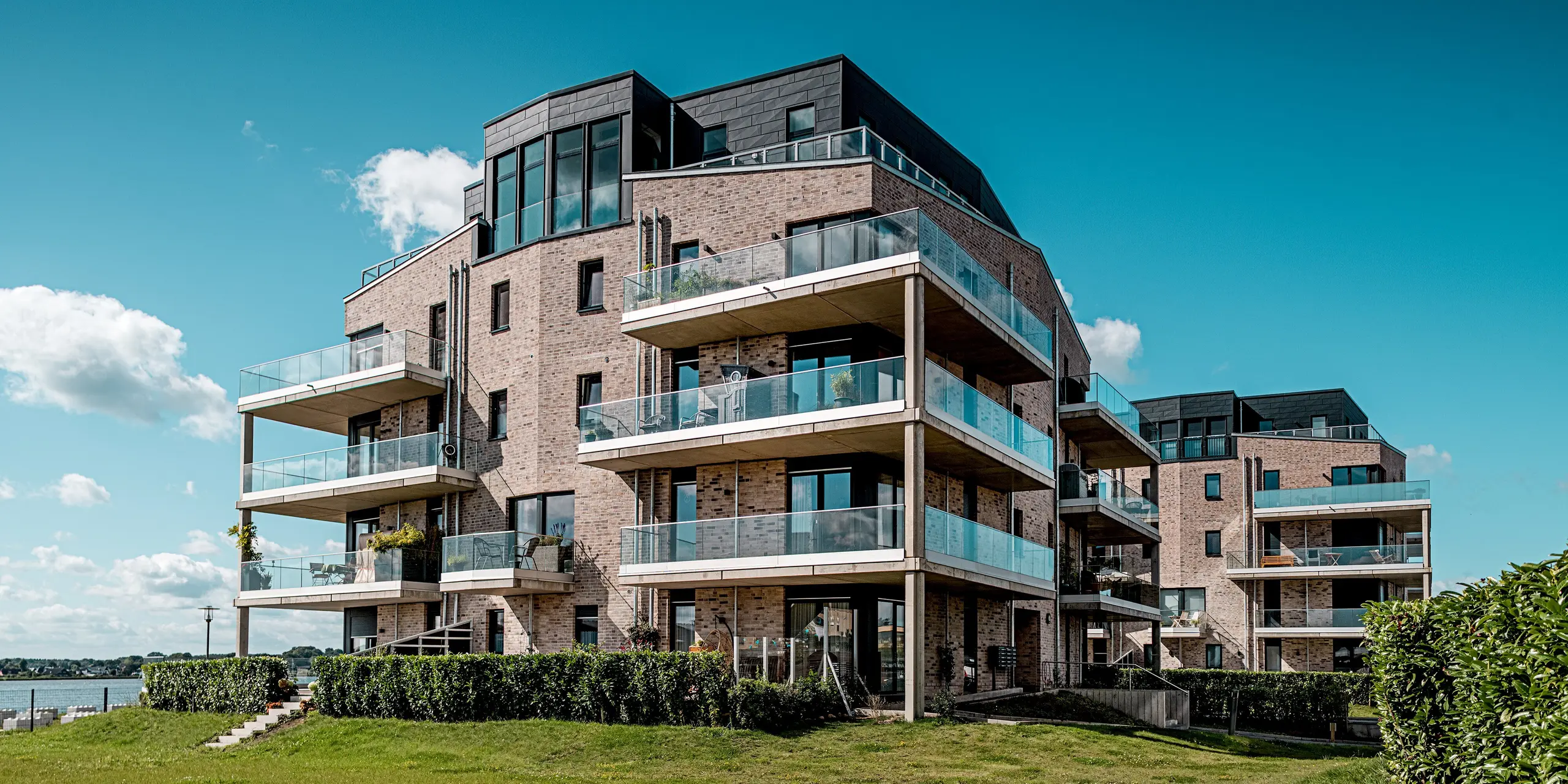 Le complexe résidentiel « Hafen zur Freiheit » (Port vers la liberté) à Schleswig présente une architecture moderne en bord de mer : des façades en briques claires côtoient des éléments Siding.X PREFA P.10 anthracite à la structure dynamique. Les étages supérieurs, particulièrement remarquables, se distinguent par leurs généreuses balustrades en verre et leurs lignes épurées qui créent un élégant contraste sous la lumière du soleil. Les façades en aluminium résistant assurent une protection durable contre les intempéries côtières et confèrent au complexe un caractère distinctif et haut de gamme. Les balcons spacieux offrent une vue imprenable sur le paysage maritime et contribuent à un confort de vie optimal.