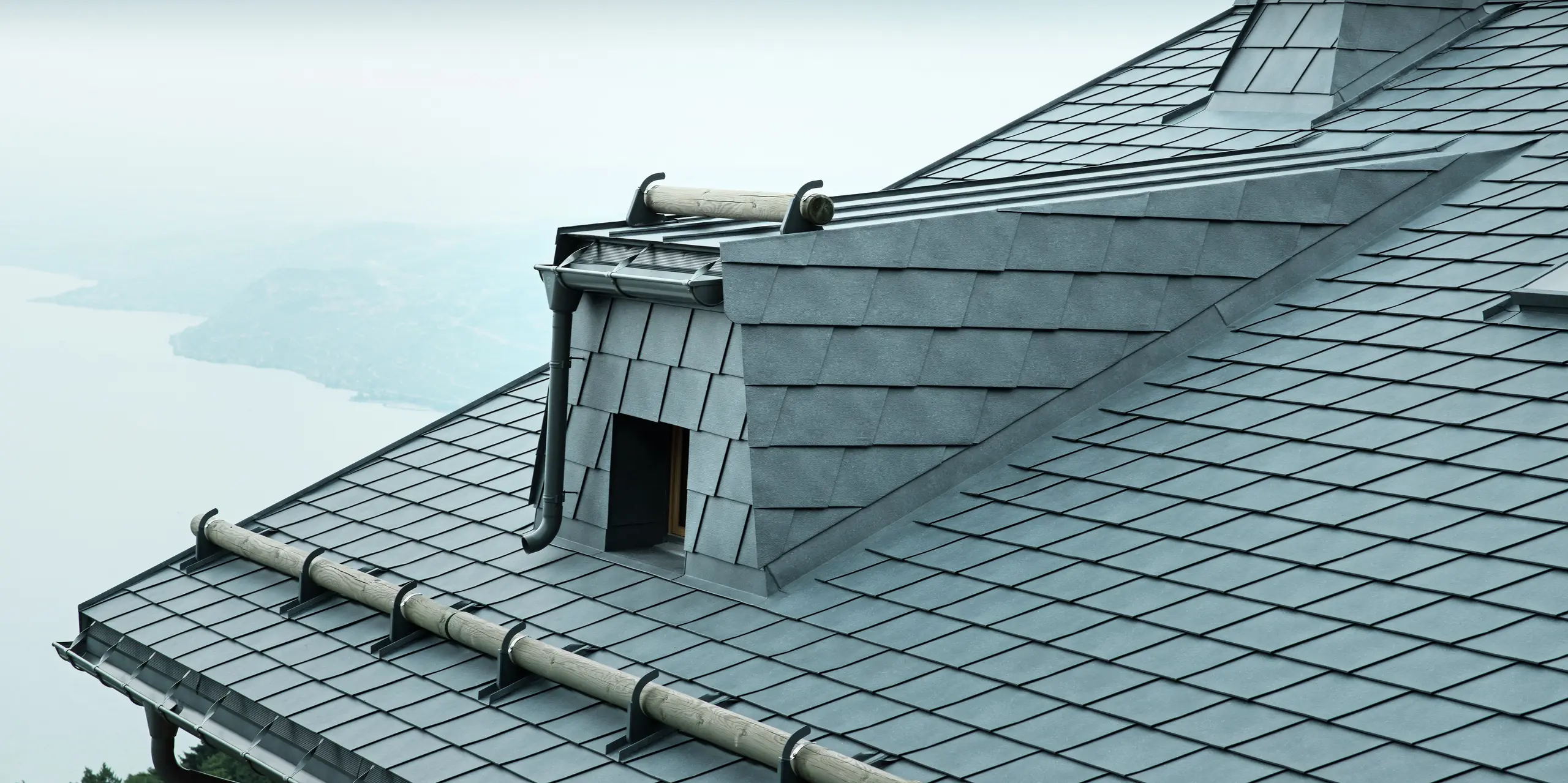 Die Dachansicht dieser modernen Holz-Lodge in der Schweiz zeigt steingraue PREFA Aluminium-Dachschindeln und eine große Dachgaube. Das langlebige Aluminiumdach schützt das Gebäude vor den extremen Wetterbedingungen am Genfersee. Der raffinierte Schneefang aus Rundholz und der markante Kamin betonen das einzigartige Design der Architektur.