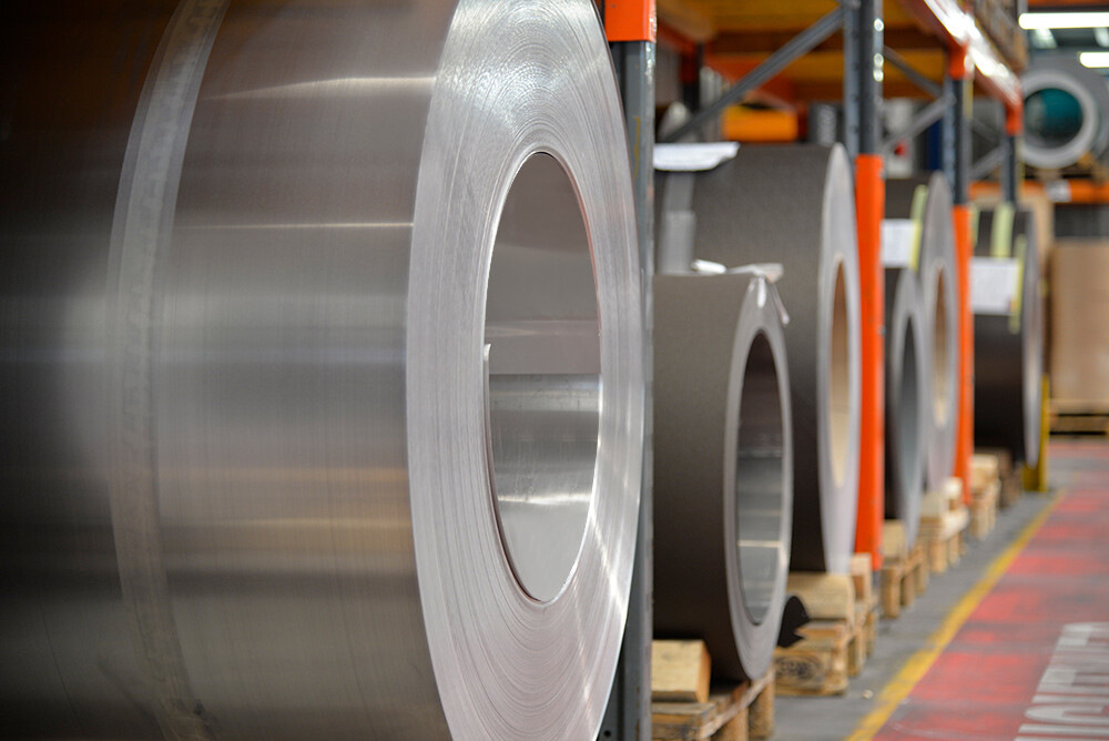 Bobines d’aluminium ("coils") sur palettes en bois : au premier plan, un rouleau en aluminium naturel et derrière, d’autres bandes et feuilles d’aluminium.