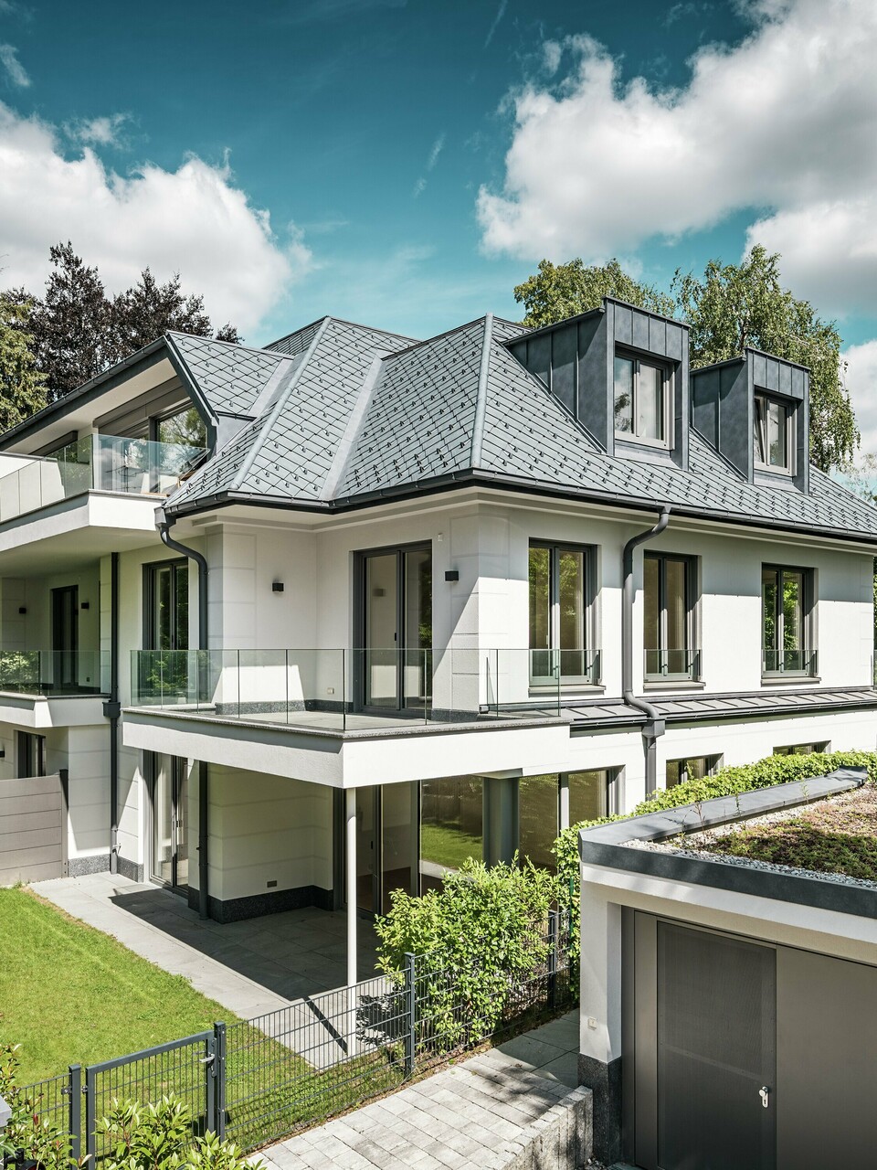 Ansicht eines modernen Mehrfamilienhauses in der Nähe von München mit stilvollem Design. Das Dach wurde mit PREFA Dachrauten 29 × 29 in P.10 Steingrau gedeckt. Die markante Dachkonstruktion wird durch elegante Flachdachgauben aus PREFALZ akzentuiert, die sowohl ästhetisch als auch funktional überzeugen. Der gepflegte Gartenbereich sowie die großzügige Terrasse betonen die hochwertige Architektur und schaffen eine harmonische Verbindung zwischen Innen- und Außenbereich. Die langlebigen und witterungsbeständigen PREFA-Produkte garantieren eine perfekte Integration in die grüne Umgebung und bieten dauerhaft Schutz und Schönheit.