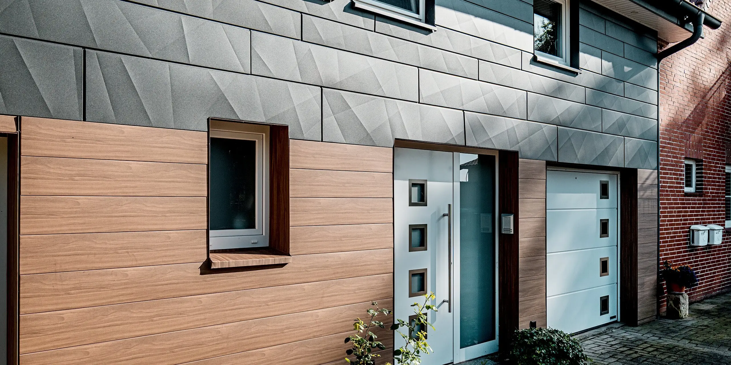 Vue détaillée de l'entrée d'une maison individuelle rénovée, dotée d'une façade moderne en aluminium PREFA. Au premier plan, le bardage PREFA noyer foncé séduit par son aspect bois de haute qualité, conférant chaleur et convivialité au rez-de-chaussée et à l'entrée. L'entrée de la maison, avec sa porte blanche et ses éléments en verre clair, est parfaitement encadrée et apporte une touche d'élégance. Au-dessus, le bardage PREFA Siding.X P.10 gris sombre, aux arêtes marquées, crée une structure de façade dynamique et de captivants jeux d'ombre et de lumière. Durables, faciles d'entretien et résistants aux intempéries, les panneaux en aluminium allient design contemporain, fonctionnalité durable et haute qualité architecturale.