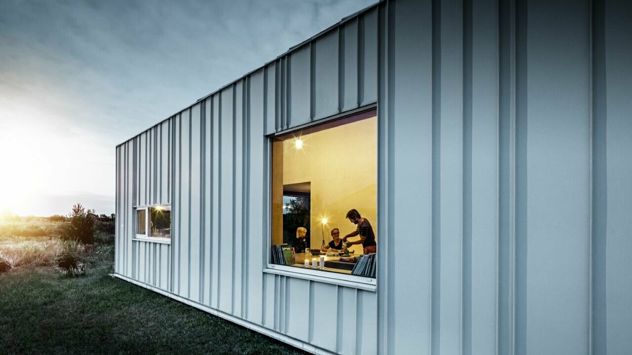 Façade avec joint debout et différentes largeurs de lames en PREFALZ de P.10 blanc PREFA photographiée au crépuscule. À travers la fenêtre de la salle à manger, on peut voir une famille en train de dîner.