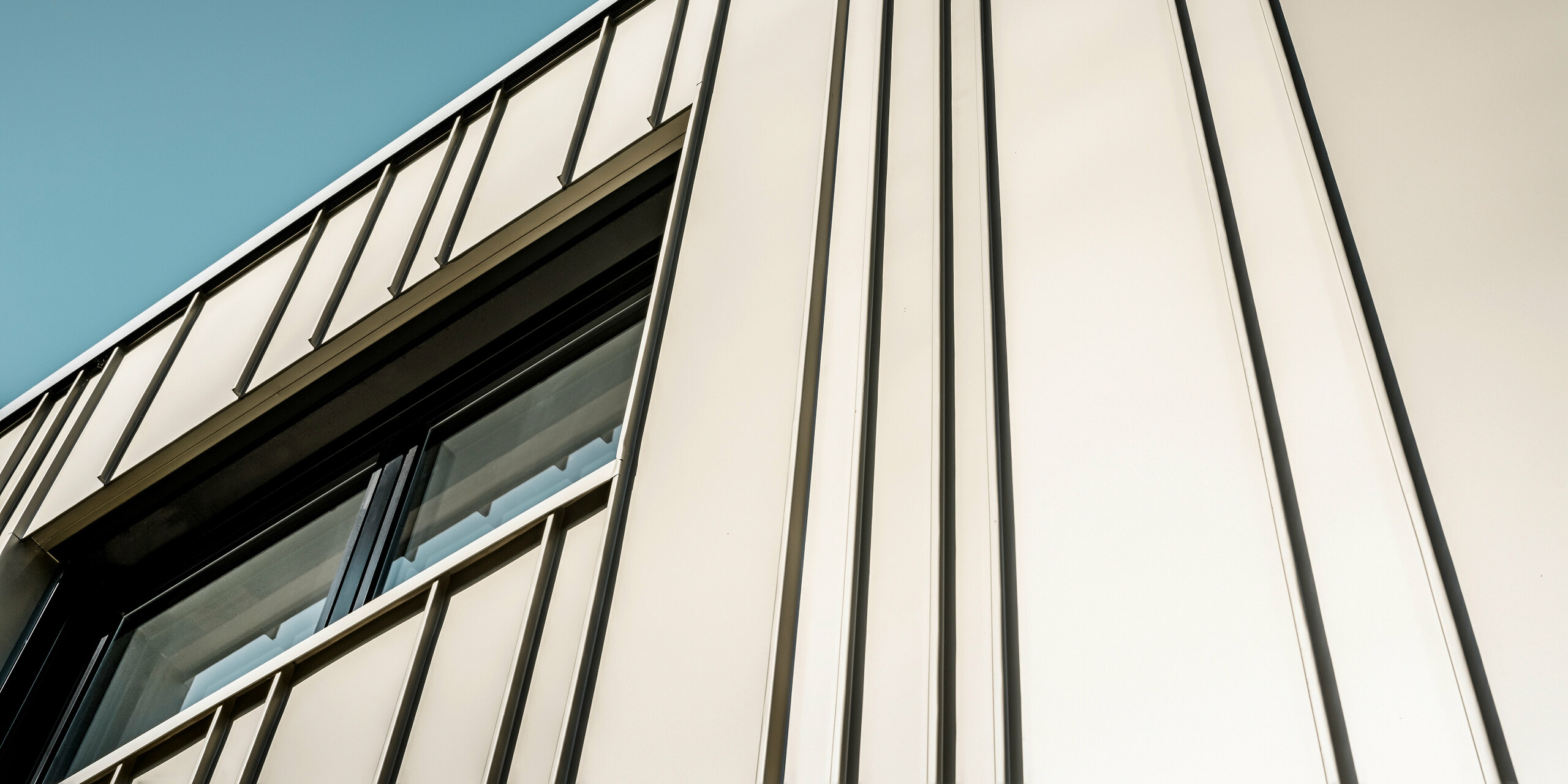 Nahaufnahme einer modernen PREFA Aluminiumfassade in Lichtbronze. Für die Winkelstehfalzfassade wurde FALZONAL in unterschiedlichen Scharenbreiten verlegt. Die Schatten der vertikalen Linien, die durch die Winkelstehfalzdeckung entstehen, heben das anspruchsvolle Design und die Präzision der Verarbeitung hervor. Das Fenster mit seiner schmalen, anthrazitfarbenen Einfassung fügt sich nahtlos in die Struktur der Fassade ein und spiegelt die Kombination aus Funktionalität, moderner Ästhetik und technischer Perfektion wider.