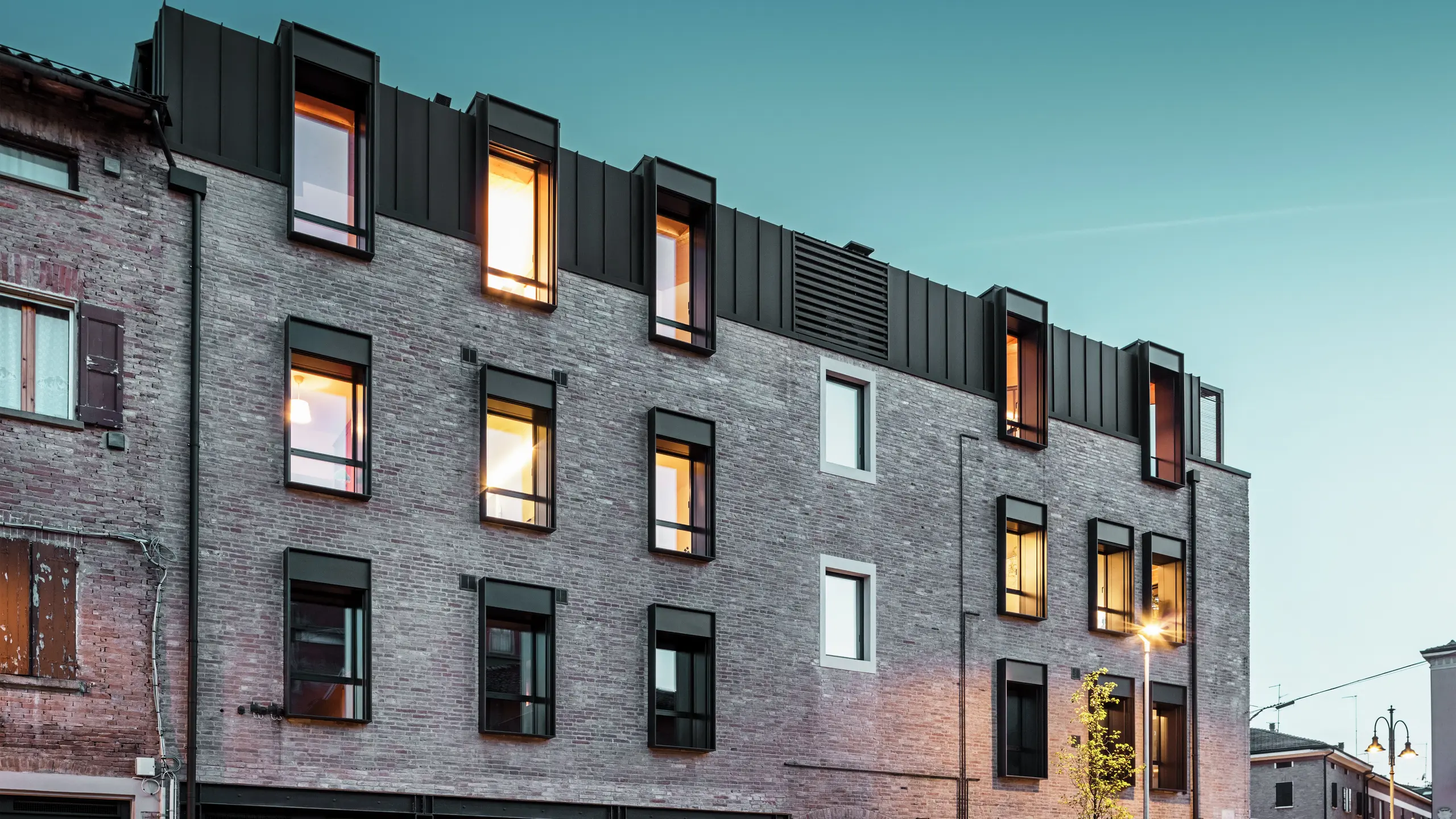 Bâtiment urbain en briques en Italie avec une toiture moderne en aluminium PREFALZ P.10 brun noisette, des fenêtres éclairées et une architecture historique environnante au crépuscule. Bâtiment urbain en briques en Italie avec toiture moderne en aluminium PREFALZ en P.10 brun noisette, fenêtres éclairées et architecture historique environnante au crépuscule.