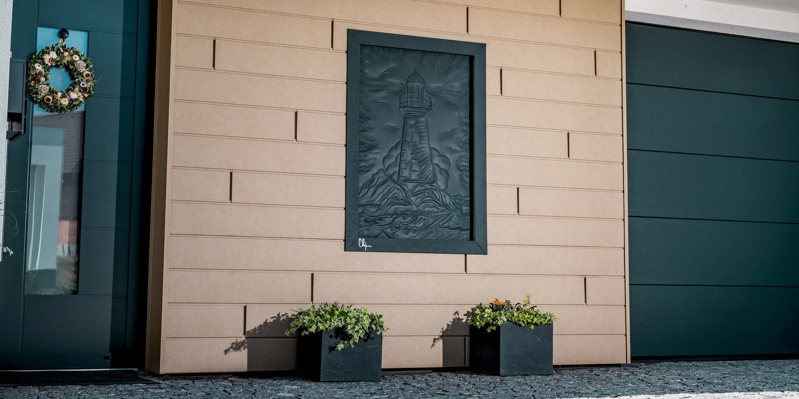 Detailaufnahme einer modernen PREFA-Sidingfassade in der Farbe P.10 Sandbraun mit kontrastierenden Aluminiumdetails in Dunkelgrau. Die langlebige Aluminiumverkleidung überzeugt durch ihre robuste Oberfläche, ihre witterungsbeständige Qualität und ihre elegante, strukturierte Optik. Direkt neben der Eingangstür befindet sich ein Eyecatcher: ein aus Aluminium gefertigtes Kunstwerk, das einen Leuchtturm im Sturm symbolisiert.