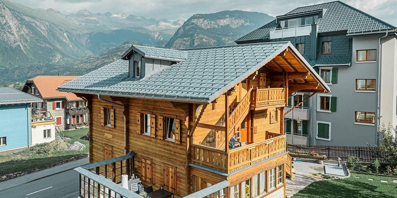 Chalet en bois suisse traditionnel avec lucarne et toit à deux pans ; la toiture a été recouverte de tuiles PREFA classiques couleur gris pierre.