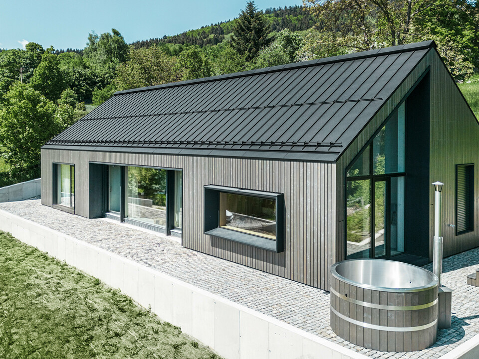 Ein modernes Holzmassivhaus in Kuncice (CZ) wurde mit großen Fensterflächen ausgestattet. Das dunkle Holzmassivhaus verfügt über ein Dachsystem aus PREFALZ in der Farbe P. 10 Schwarz. Im hinteren Außenbereich steht ein Whirlpool mit einer Holzverkleidung.