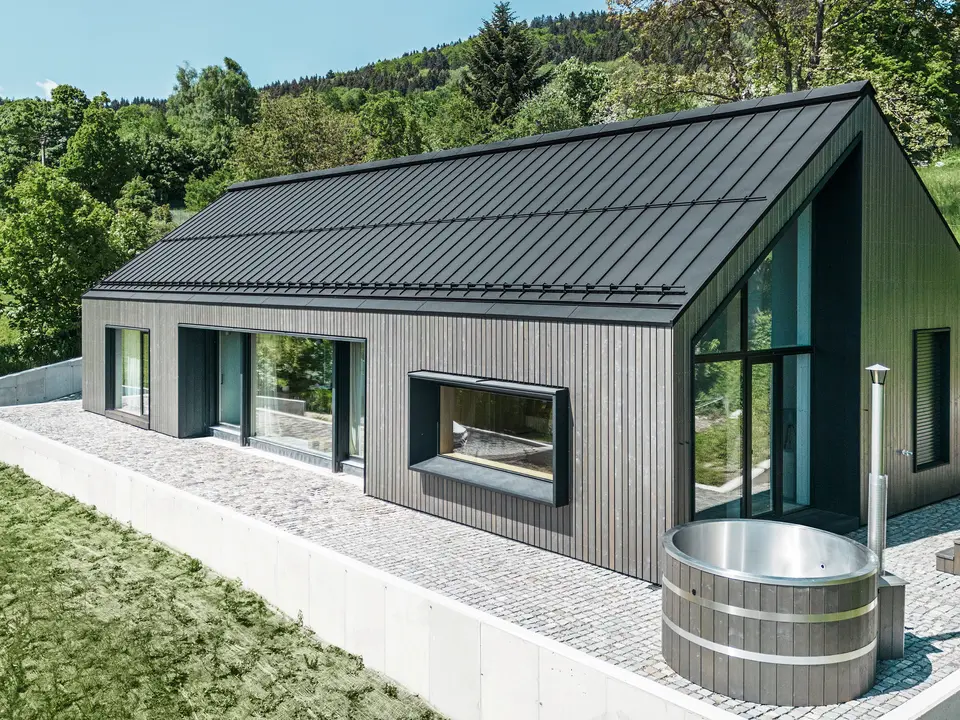 Ein modernes Holzmassivhaus in Kuncice (CZ) wurde mit großen Fensterflächen ausgestattet. Das dunkle Holzmassivhaus verfügt über ein Dachsystem aus PREFALZ in der Farbe P. 10 Schwarz. Im hinteren Außenbereich steht ein Whirlpool mit einer Holzverkleidung.