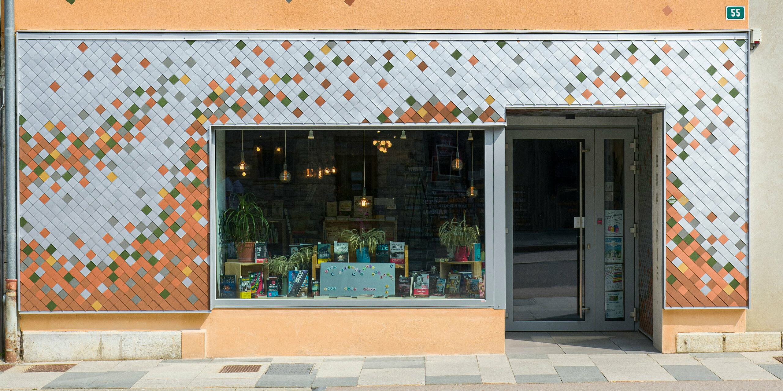 Vue de face de la façade colorée d'une librairie de la petite communauté française des Rousses. Le bâtiment combine un étage supérieur orange vif avec des éléments décoratifs et un étage inférieur avec une façade en forme de mosaïque en aluminium de différentes couleurs. Une grande fenêtre au rez-de-chaussée présente des livres et des plantes à côté de la porte d'entrée. Une étroite gouttière longe le côté du bâtiment et le numéro 55 est visible sur un panneau vert à côté de la porte.