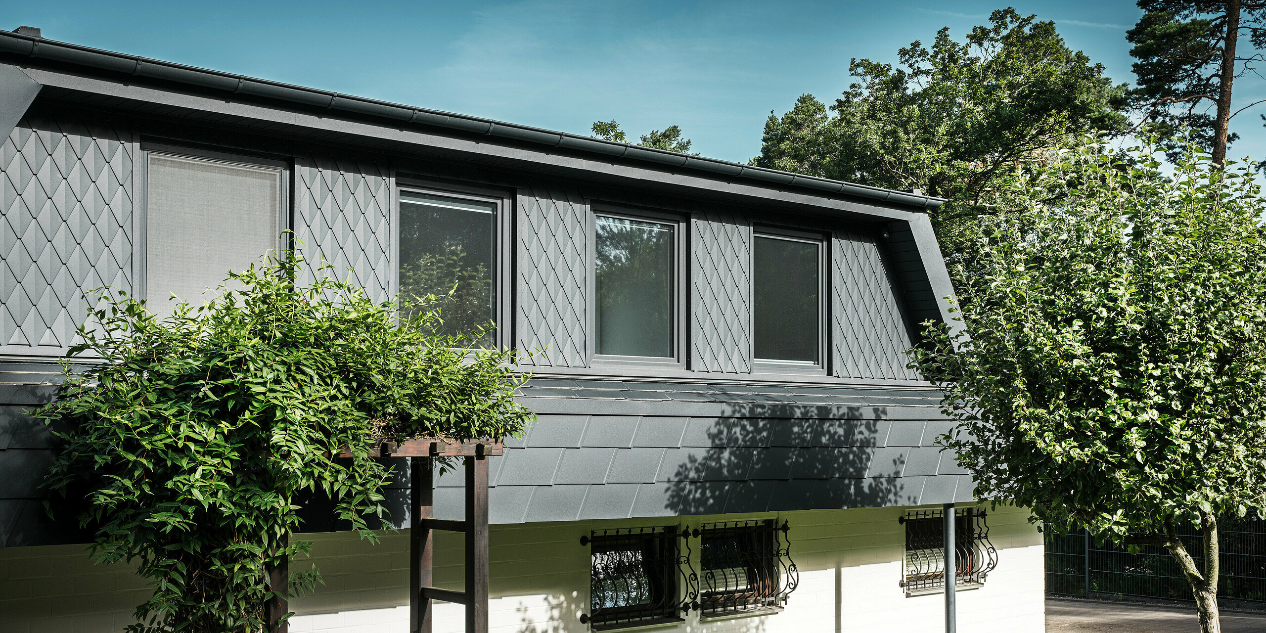 Vue latérale d'une maison individuelle à Mechernich avec de grandes lucarnes trapézoïdales. Les lucarnes sont équipées de petits losanges PREFA en P.10 anthracite, tandis que le toit est recouvert des DS.19 PREFA, également en P.10 anthracite, qui ont fait leurs preuves. Les losanges et bardeaux de toiture en aluminium offrent une solution résistante aux intempéries, esthétique et durable pour la toiture et la façade. La maison unifamiliale traditionnelle est entourée d'un jardin bien entretenu et de nombreux arbres.