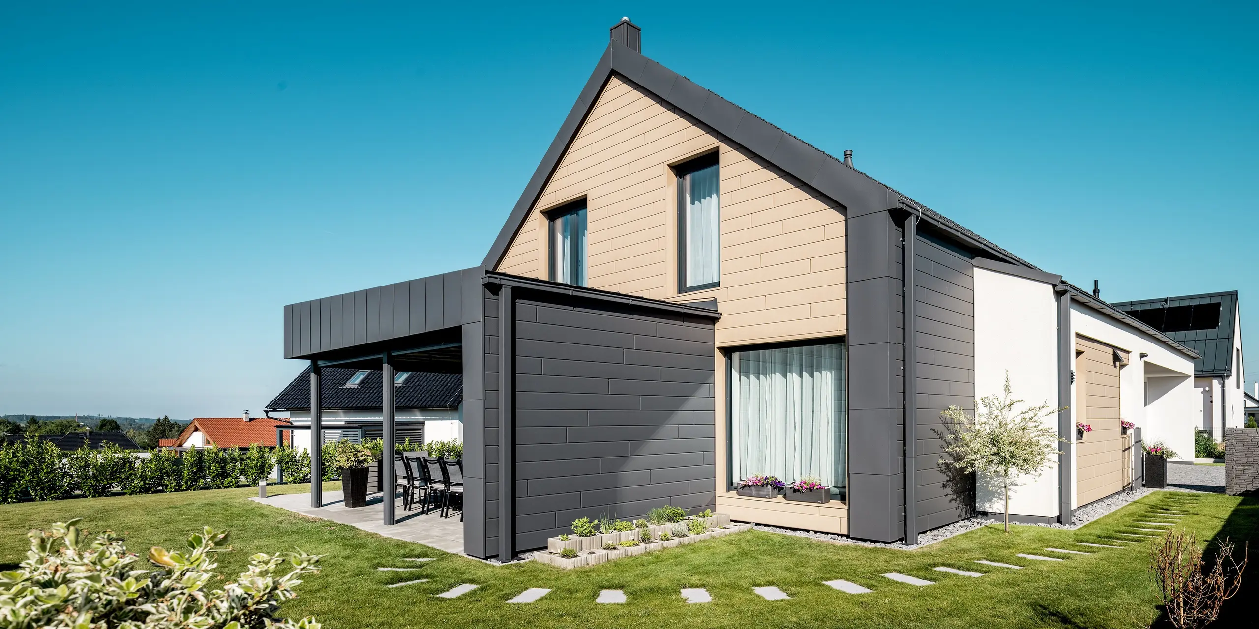 Modernes Einfamilienhaus mit harmonisch abgestimmter PREFA Aluminium-Gebäudehülle. Die Kombination aus PREFA Sidings in P.10 Dunkelgrau und P.10 Sandbraun sowie den klaren Dachflächen schafft eine elegante Verbindung aus Kontrast und Einheit. Die großzügige Gartenansicht zeigt die zeitgemäße Architektur mit langlebigen, witterungsbeständigen Aluminiumoberflächen, die Schutz und stilvolles Design vereinen.