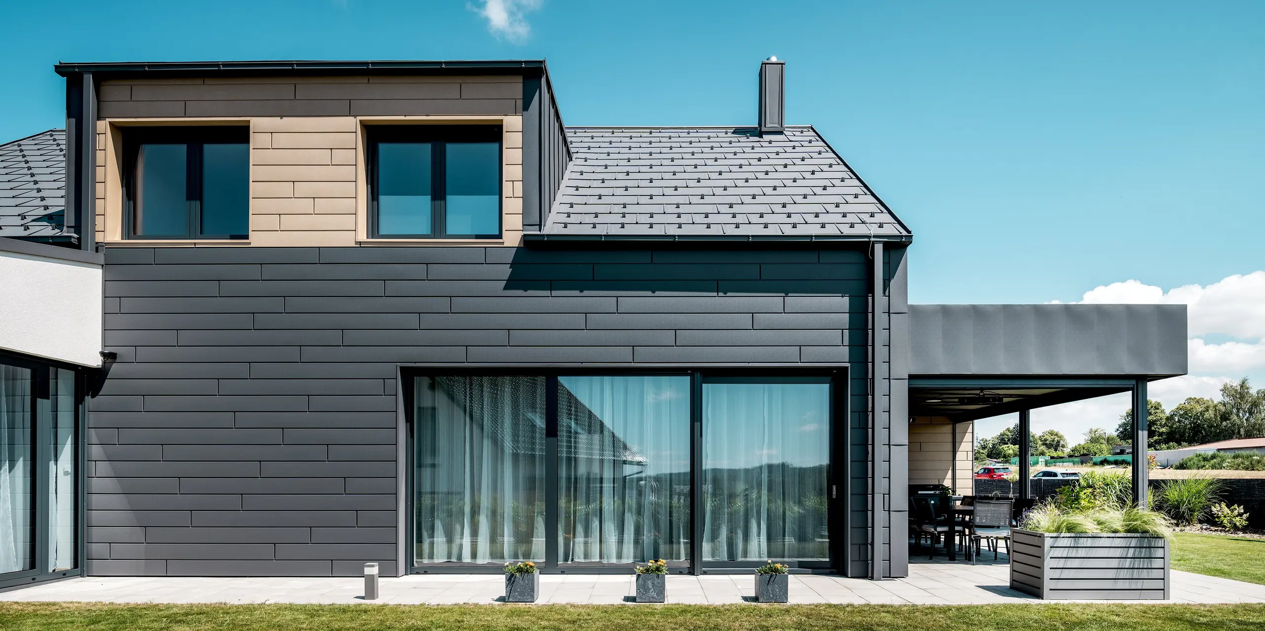 Modernes Einfamilienhaus mit klarer Architektur und harmonischer Aluminiumgestaltung. Die großflächige Verglasung öffnet den Wohnbereich zum Garten, während die dunkle PREFA Siding-Fassade und das Dach in P.10 Dunkelgrau eine elegante, zeitlose Optik schaffen. Die Kombination aus geradlinigen Formen, heller Umgebung und hochwertigen Aluminiumoberflächen zeigt eine moderne Gebäudehülle, die durch Langlebigkeit, Widerstandsfähigkeit und stilvolle Details überzeugt.