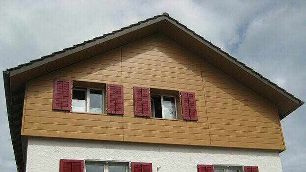Fassadensanierung mit Sidings von PREFA Giebelverkleidung eines klassischen Hauses mit Satteldach. Der Giebel ist mit horizontal verlegten PREFA Sidings in Eiche natur verkleidet. Die Fenster haben rote Fensterläden.
