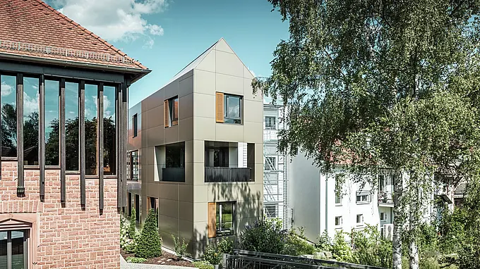 La photo montre la mairie moderne d'Alzenau, dont la façade est composée de panneaux composites en aluminium PREFA de couleur bronze. Le toit pointu et les fenêtres clairement structurées avec des stores en bois confèrent au bâtiment une esthétique élégante et contemporaine. Au premier plan, on aperçoit un espace vert avec un chemin pavé et des arbres qui complète harmonieusement le design urbain du bâtiment.