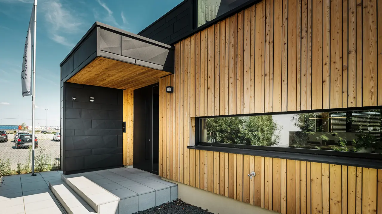 Entrée latérale de la maison modèle Autark du centre de maisons préfabriquées Blaue Lagune à Vösendorf, avec une façade moderne en bois naturel et des panneaux PREFA Siding.X noirs. L'image montre la combinaison élégante du revêtement en bois et de l'aluminium dans le revêtement de haute qualité P.10 noir.