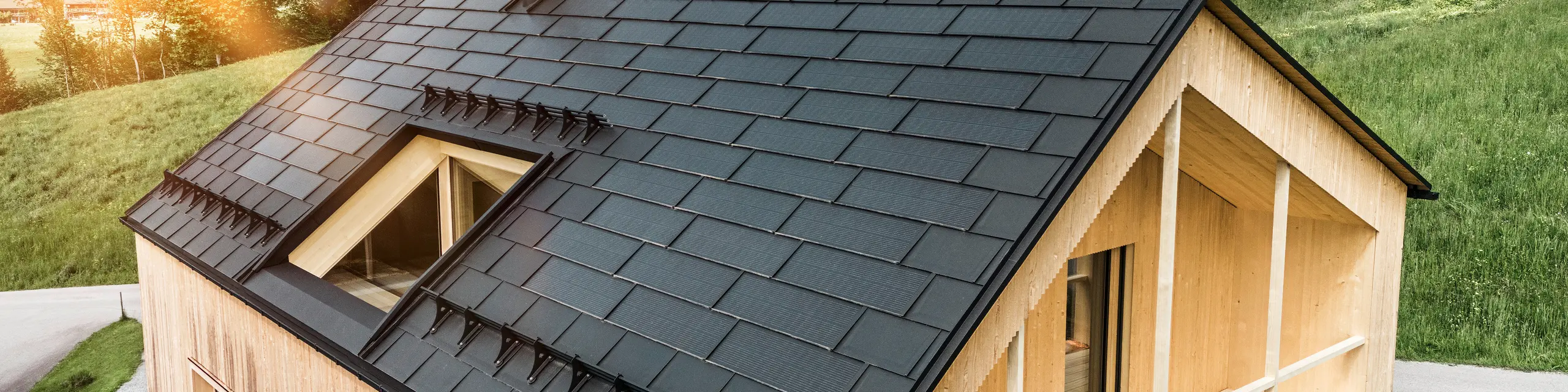 Blick auf ein modernes, nachhaltiges Holzhaus in Egg mit einem PREFA Solardach, das durch seine dunkle, strukturierte sowie elegante Erscheinung auffällt. Die Solarmodule sind strategisch zwischen den klassischen Dachplatten R.16 platziert, um eine optimale Sonneneinstrahlung zu nutzen und sich dabei ästhetisch in das Gesamtdesign einzufügen. Das Haus steht im Einklang mit seiner grünen Umgebung, während die warme Abendsonne die natürliche Holztextur der Fassade unterstreicht und die solide Bauweise betont. Ein harmonisches Beispiel für umweltbewusstes Bauen und Energieeffizienz.