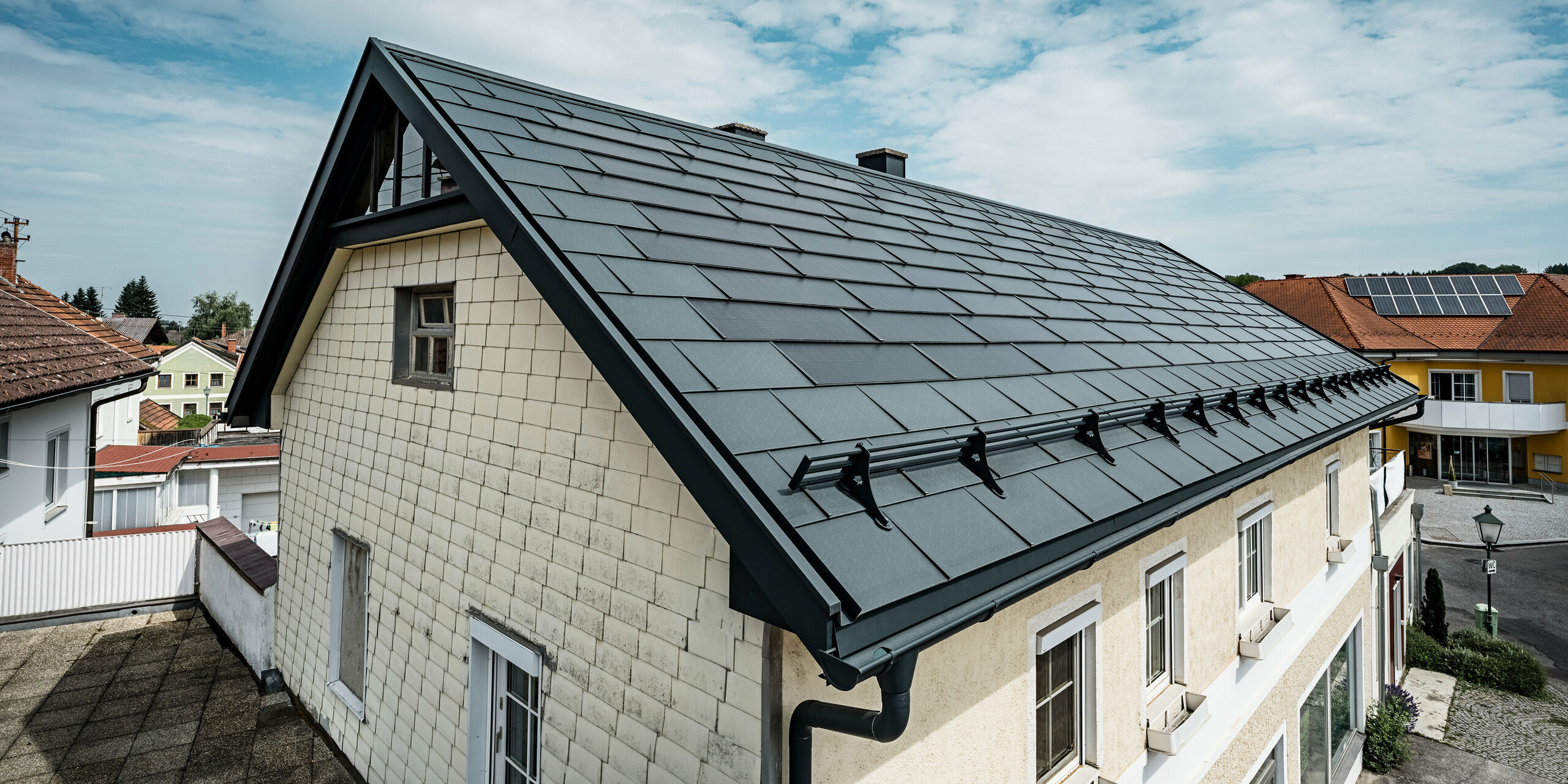 Vue plongeante sur le toit d'une maison familiale à Mettmach, en Autriche. Dans le cadre d'une rénovation, la maison a été équipée de tuiles solaires PREFA en anthracite. La surface structurée des tuiles s'harmonise avec l'élégance simple de la maison. Un système de pare-neige est fixé au bord du toit. D'autres maisons dotées de solutions de toit traditionnelles et solaires sont visibles à l'arrière-plan, mettant en valeur la conception moderne et économe en énergie du toit dans un cadre rural.