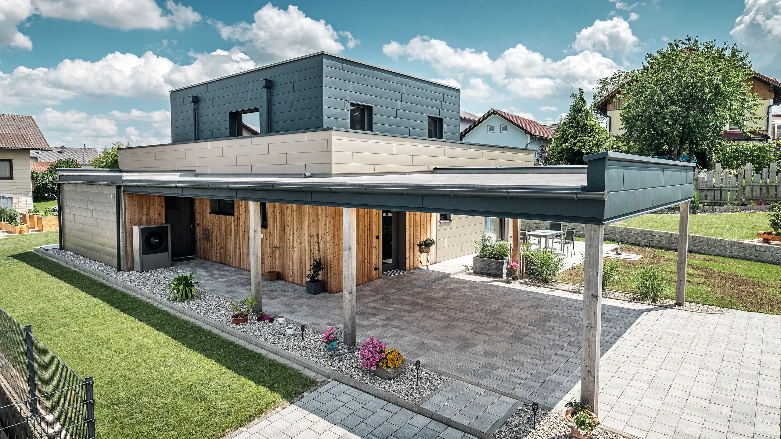 Vue aérienne de l'allée d'une maison familiale moderne à Wippenham, en Autriche, avec une façade en aluminium PREFA bicolore bronze et anthracite. La maison montre l'intégration élégante des panneaux de façade Siding.X et du revêtement en bois. Le carport couvert, parfaitement intégré à l'architecture de la maison, est clairement visible. Le jardin et la terrasse bien entretenus soulignent le design moderne et fonctionnel du bâtiment.