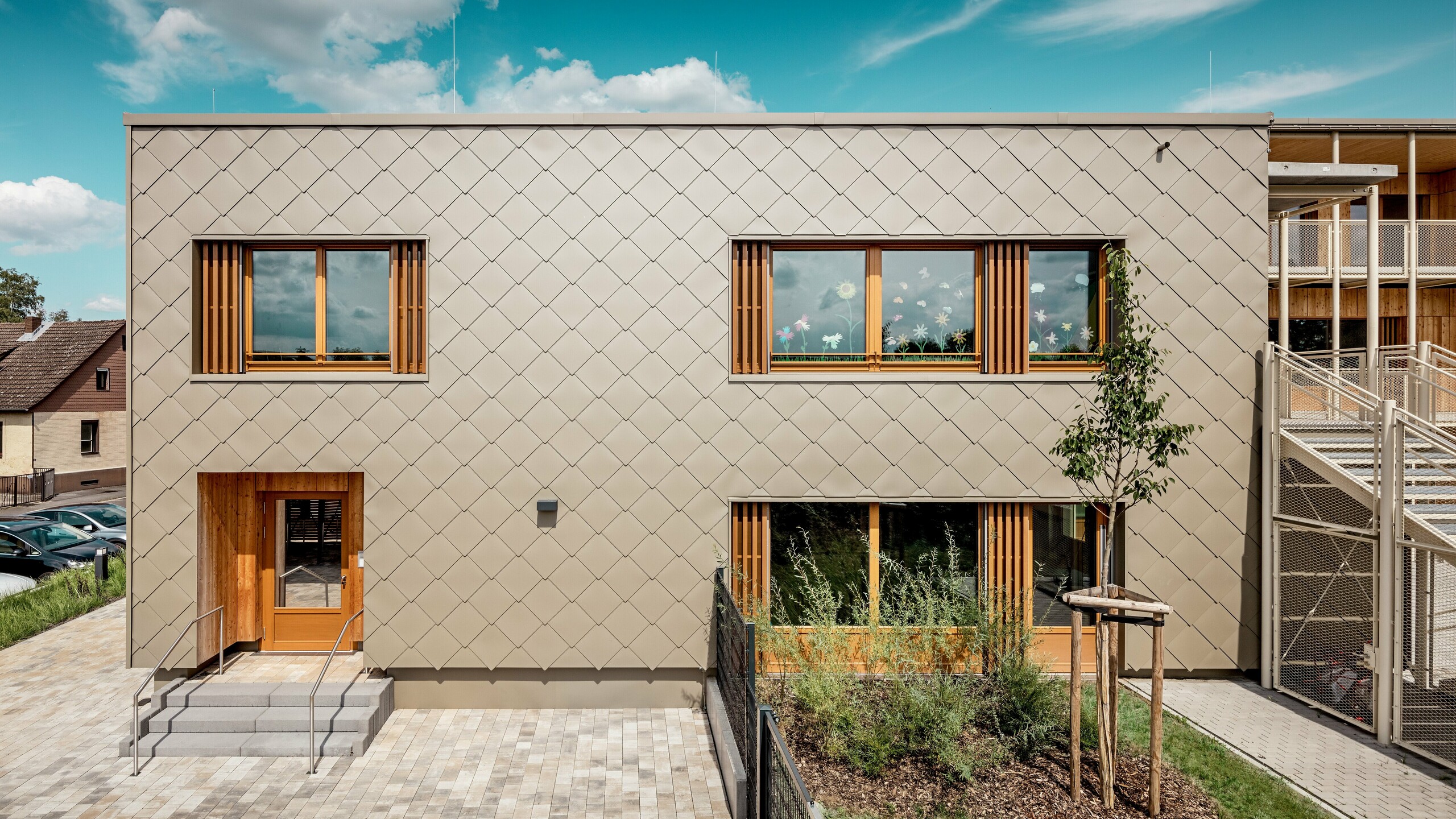 Die Rückseite der modernen Kindertagesstätte in Alzenau wird aus einer frontalen Perspektive gezeigt und beeindruckt mit klaren Linien und einer markanten Fassade aus bronzefarbenen PREFA Wandrauten 44 × 44. Die geometrische Fassadenstruktur erzeugt ein harmonisches Schuppenmuster, das durch die ruhige, horizontale Ausrichtung der Fenster akzentuiert wird. Großzügige Holzrahmen und vertikale Lamellen setzen natürliche Akzente und sorgen für eine warme, einladende Atmosphäre. Die durchdachte Kombination aus langlebigem Aluminium und regionalem Holz steht für nachhaltige Architektur mit hoher gestalterischer Qualität. Das Bild betont die präzise Ausführung, die hochwertige Materialwahl und die gelungene Integration des Gebäudes in das städtische Umfeld.