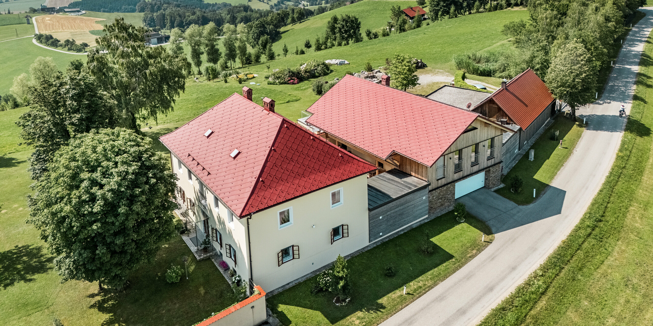 Das Bild zeigt die sanierten Einfamilienhäuser in Aschach an der Steyr, deren Dächer mit PREFA Dachrauten 44 x 44 in der Farbe P.10 Oxydrot gedeckt wurden. Die rote Farbe der Aluminiumdächer fügt sich harmonisch in die idyllische Landschaft ein und sorgt für eine markante, langlebige Optik. PREFA-Produkte garantieren Ästhetik und Widerstandsfähigkeit.