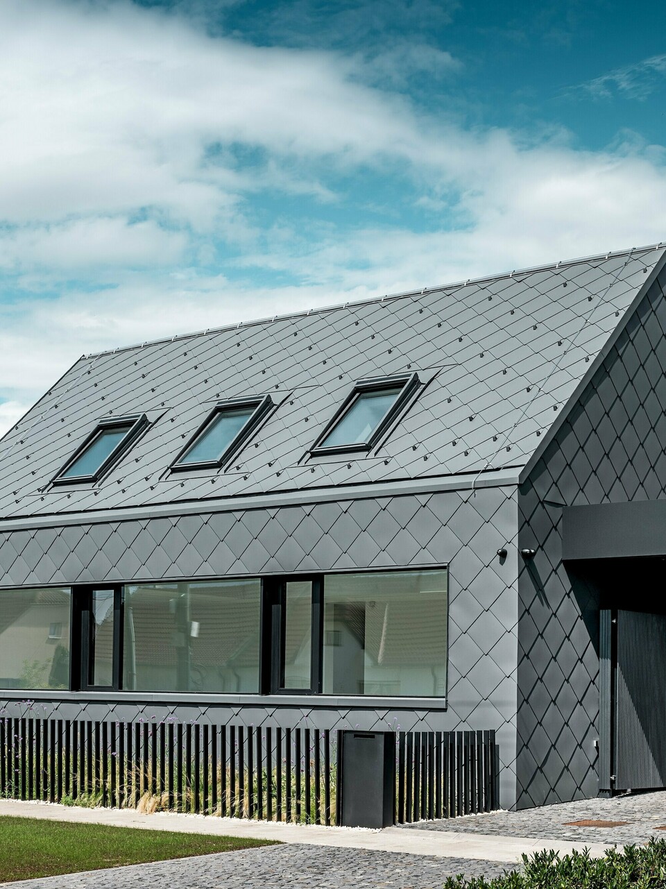 Maison individuelle moderne à Trnava-Modranka avec bardage aluminium : l’enveloppe de cette structure minimaliste est entièrement revêtue de losanges 44x44 PREFA P.10 gris sombre, utilisées pour la toiture et les façades. Le motif losange, saisissant, se déploie harmonieusement sur le toit et la façade, conférant à la maison une allure monolithique et affirmée. De larges fenêtres soulignent l’architecture, tandis que l’aluminium PREFA, résistant aux intempéries, assure une protection durable et une élégante finition mate. Le ciel lumineux accentue le contraste avec l’enveloppe sombre du bâtiment et met en valeur son design moderne.