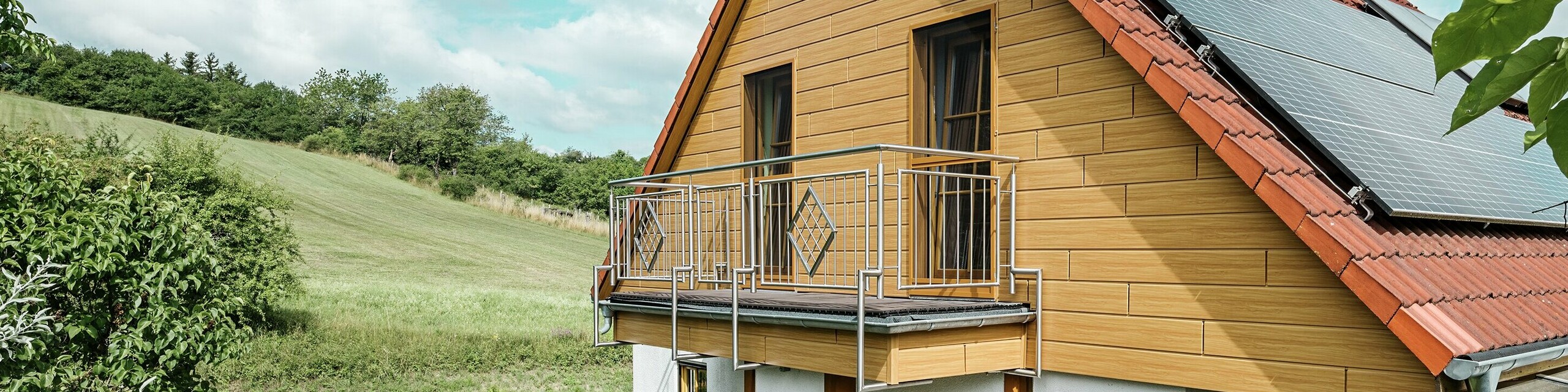 La photo montre une charmante maison individuelle à Blieskastel, dont le pignon fraîchement rénové avec des Sidings PREFA couleur chêne naturel, attire immédiatement le regard. L'aspect bois de l'aluminium, plus durable, s'harmonise parfaitement avec le toit en tuiles rouges et la vaste véranda. La verdure luxuriante et le parterre de fleurs soigneusement aménagé forment une scène idyllique et vivante. La rénovation de façades avec PREFA confère à la maison un aspect naturel et en impose par sa robustesse : résistance aux intempéries, fidélité aux couleurs d'origine et longue durée de vie, pratiquement sans entretien.