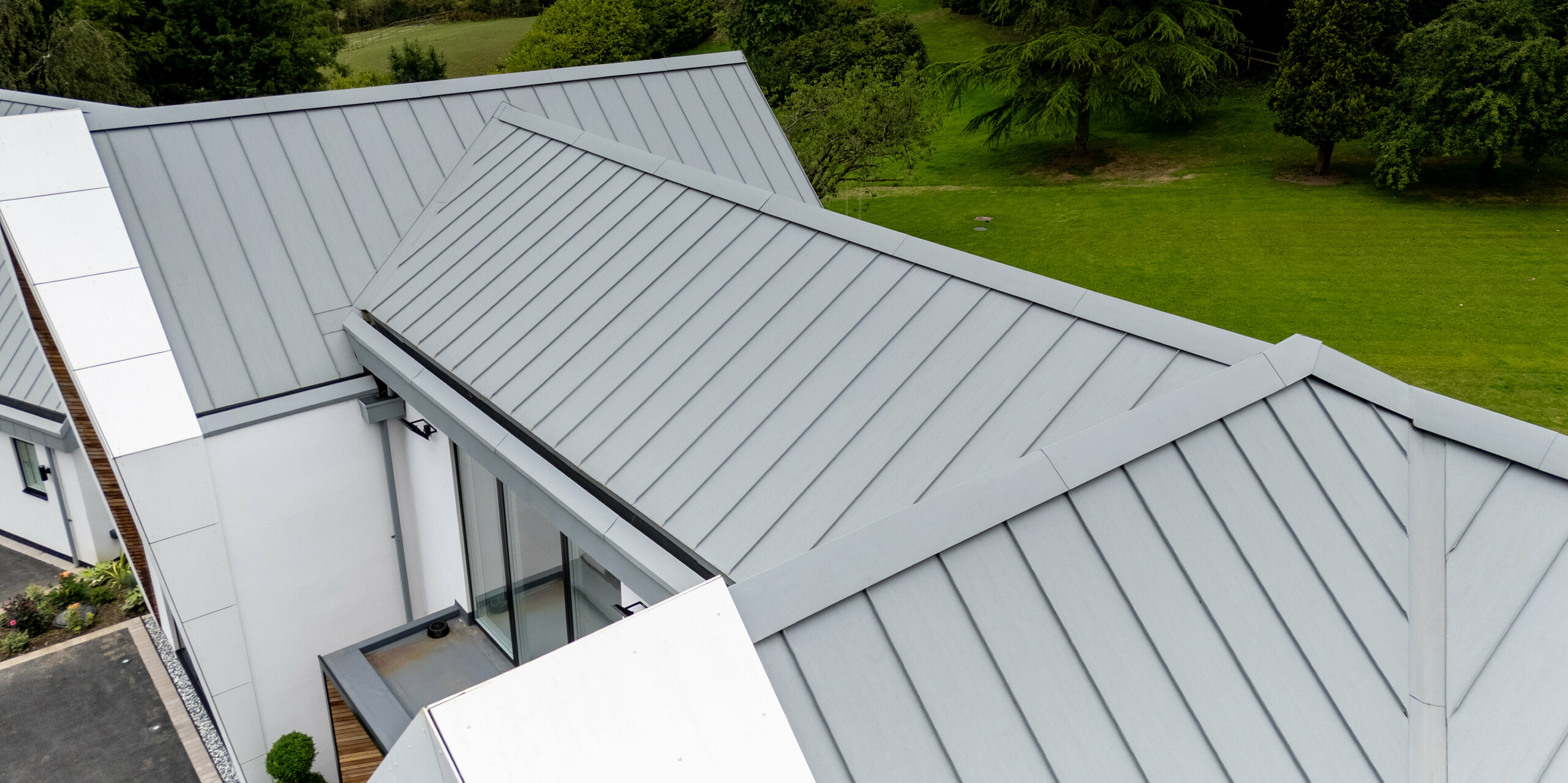 Die Aufnahme zeigt den First des präzise ausgeführten PREFALZ-Dachs in der Farbe Patinagrau aus einer einzigartigen Perspektive. Die Stehfalzdeckung mit ihren klaren Linien und der eleganten Metalloberfläche hebt sich hervorragend von der grünen Umgebung ab. Durch den Einsatz von PREFALZ sind nicht nur eine ansprechende Optik, sondern auch Langlebigkeit und Witterungsbeständigkeit gewährleistet. Die perfekte Integration in das moderne Design des Gebäudes zeigt eindrucksvoll, wie Funktion und Form miteinander verschmelzen.