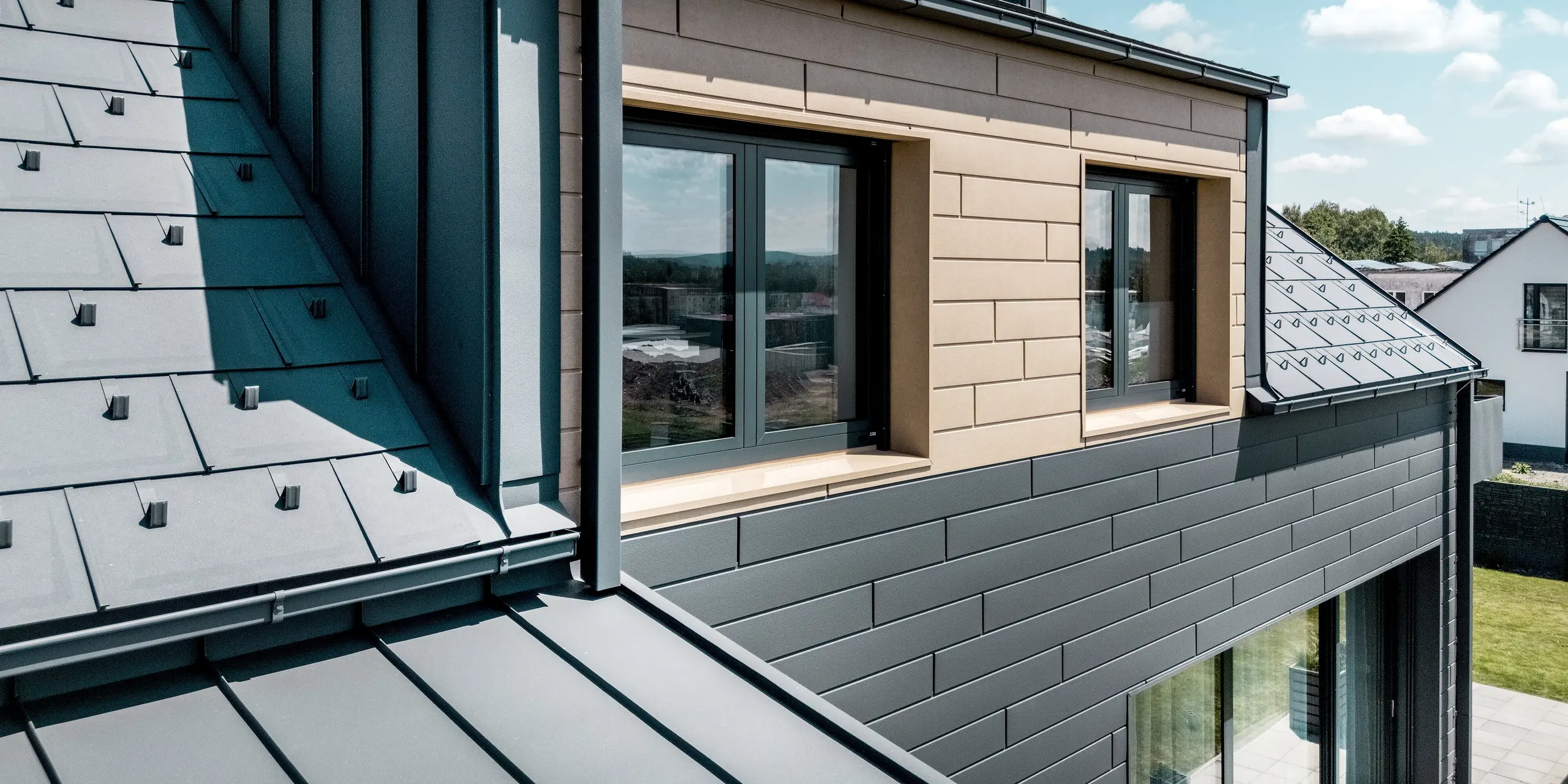 Diese Detailansicht zeigt die Flachdachgaube eines Einfamilienhauses mit einer modernen PREFA Aluminiumfassade. Die Kombination aus PREFA Sidings in den Farben P.10 Dunkelgrau und P.10 Sandbraun sowie einem Dach aus PREFA Dachplatten R.16 und PREFALZ zeigt die präzise Abstimmung von Materialien und Farben. Die klaren Linien der Gebäudehülle verbinden zeitgemäßes Design mit den Vorteilen von Aluminium: Es ist langlebig, witterungsbeständig und pflegeleicht.