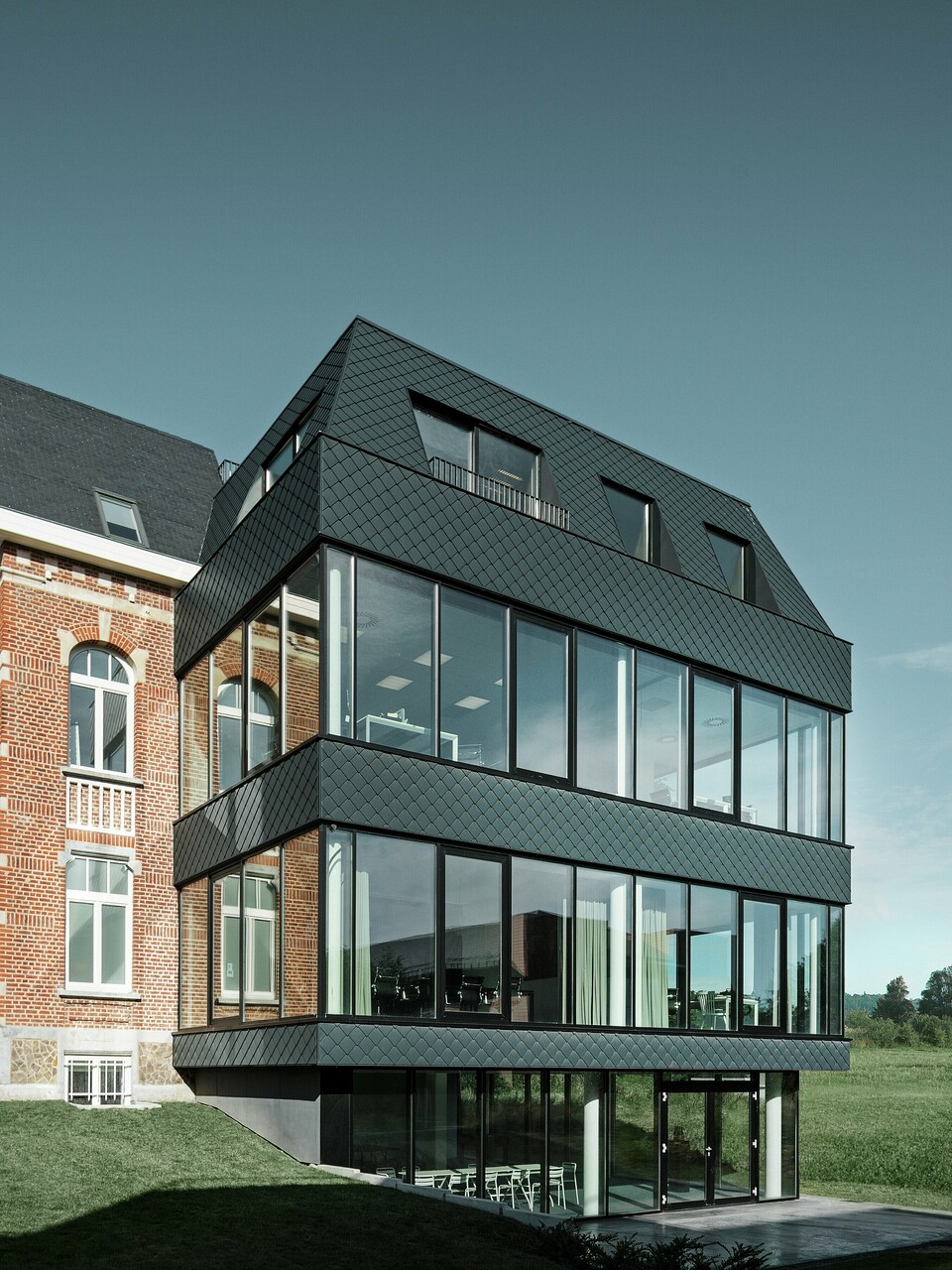 Seitlicher Blick auf das Kasteeltje Heizel, wo historischer Backsteinbau mit Turm nahtlos in einen modernen Anbau übergeht. Die anthrazitfarbenen PREFA Wandrauten setzen mit ihrer markanten Struktur elegante Kontraste zu den großzügigen Glasflächen. Die klare Linienführung des Neubaus verstärkt die Verbindung von Tradition und moderner Architektur in einer eindrucksvollen Kulisse.