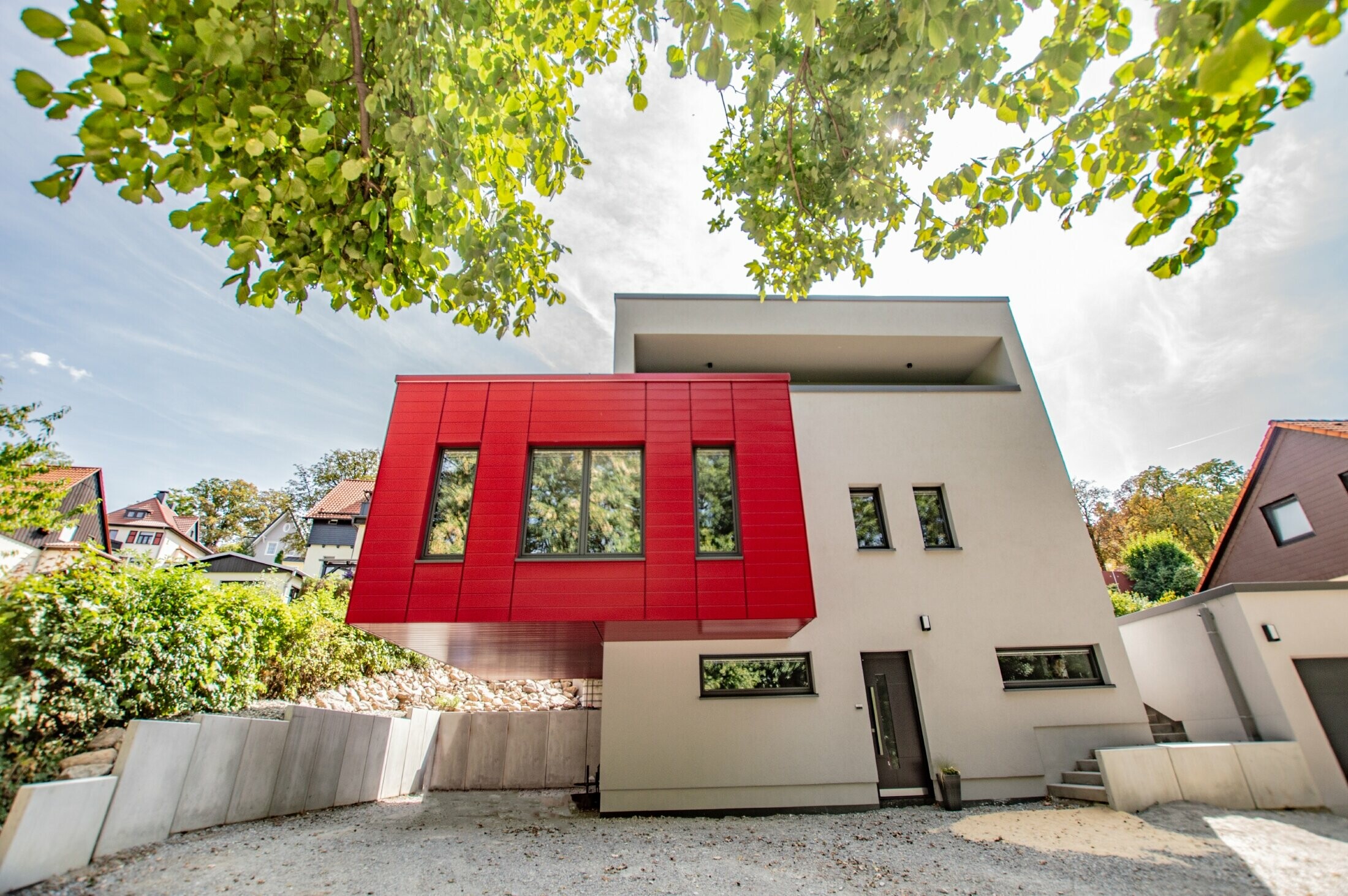 Partie de bâtiment en porte-à-faux recouverte de Sidings PREFA couleur rouge rubis. Grand oriel rouge qui détonne par rapport à la façade blanche.