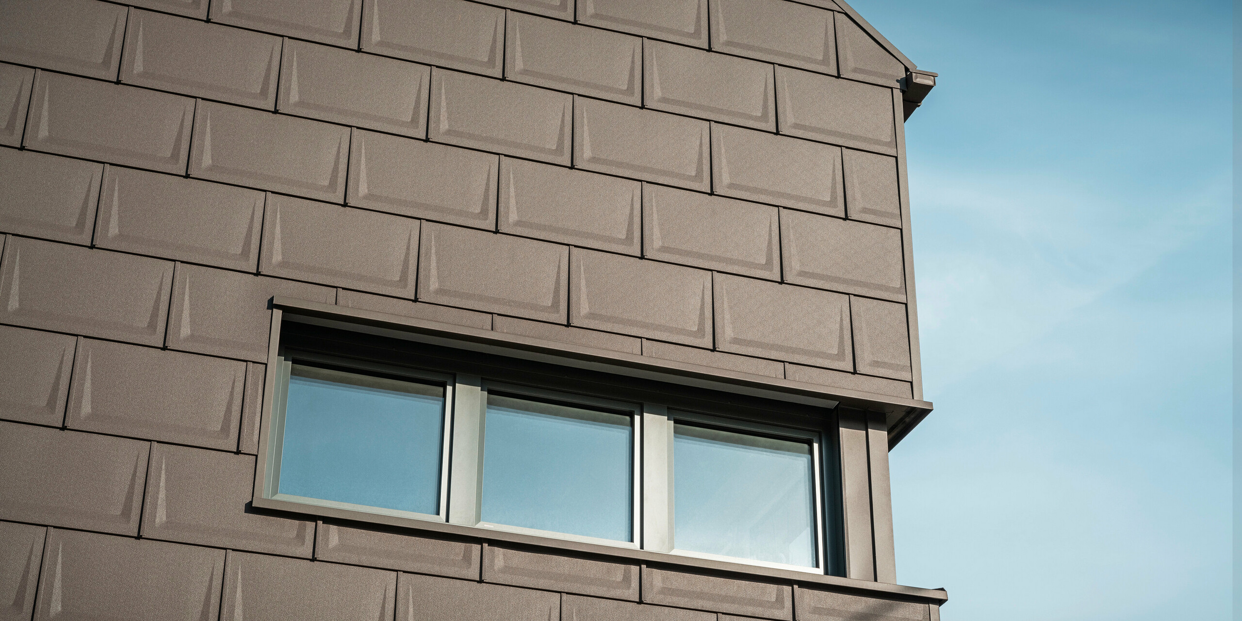 Nahaufnahme der Fassade eines Einfamilienhauses in Neukirchen, Österreich, gestaltet mit der innovativen PREFA Dachplatte R.16 in P.10 Braun, die als objektbezogene Sonderlösung sowohl für das Dach als auch für die Fassadengestaltung des Obergeschosses verwendet wird. Diese einzigartige Anwendung sorgt für eine harmonische und ästhetische Optik und unterstreicht die Vielseitigkeit der PREFA Aluminiumprodukte. Die einheitliche Farbgebung und das durchdachte Designkonzept des Gebäudes betonen die vielseitigen Gestaltungsmöglichkeiten von PREFA.