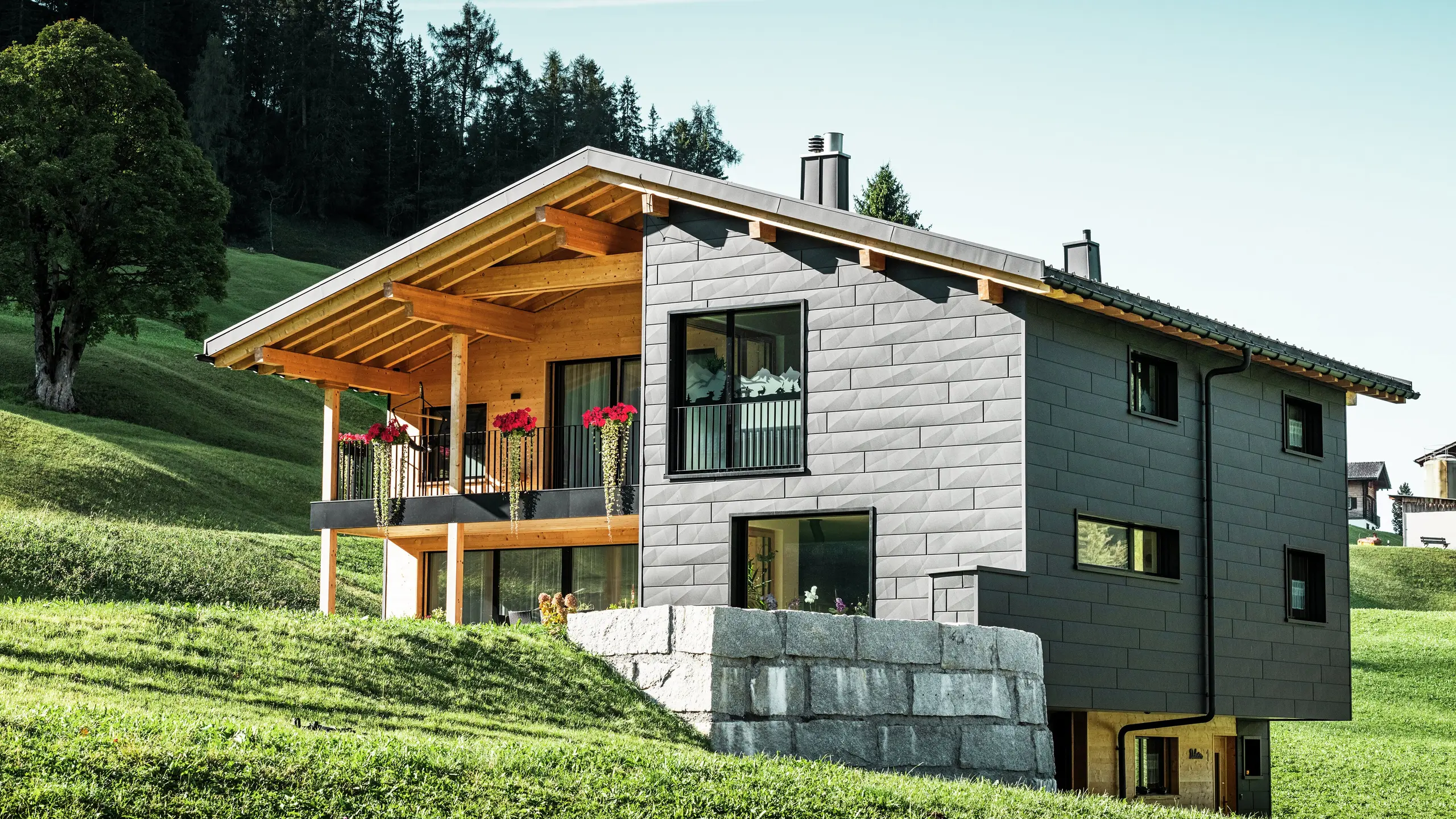 Cette maison individuelle moderne à Ascharina, en Suisse, s'intègre harmonieusement dans le paysage alpin verdoyant. Le bâtiment est entièrement revêtu de panneaux de façade PREFA Siding.X durables et résistants aux intempéries en P.10 noir, qui confèrent à la structure une apparence unique. Le balcon en bois complète parfaitement la coque en aluminium foncé et apporte une touche naturelle.