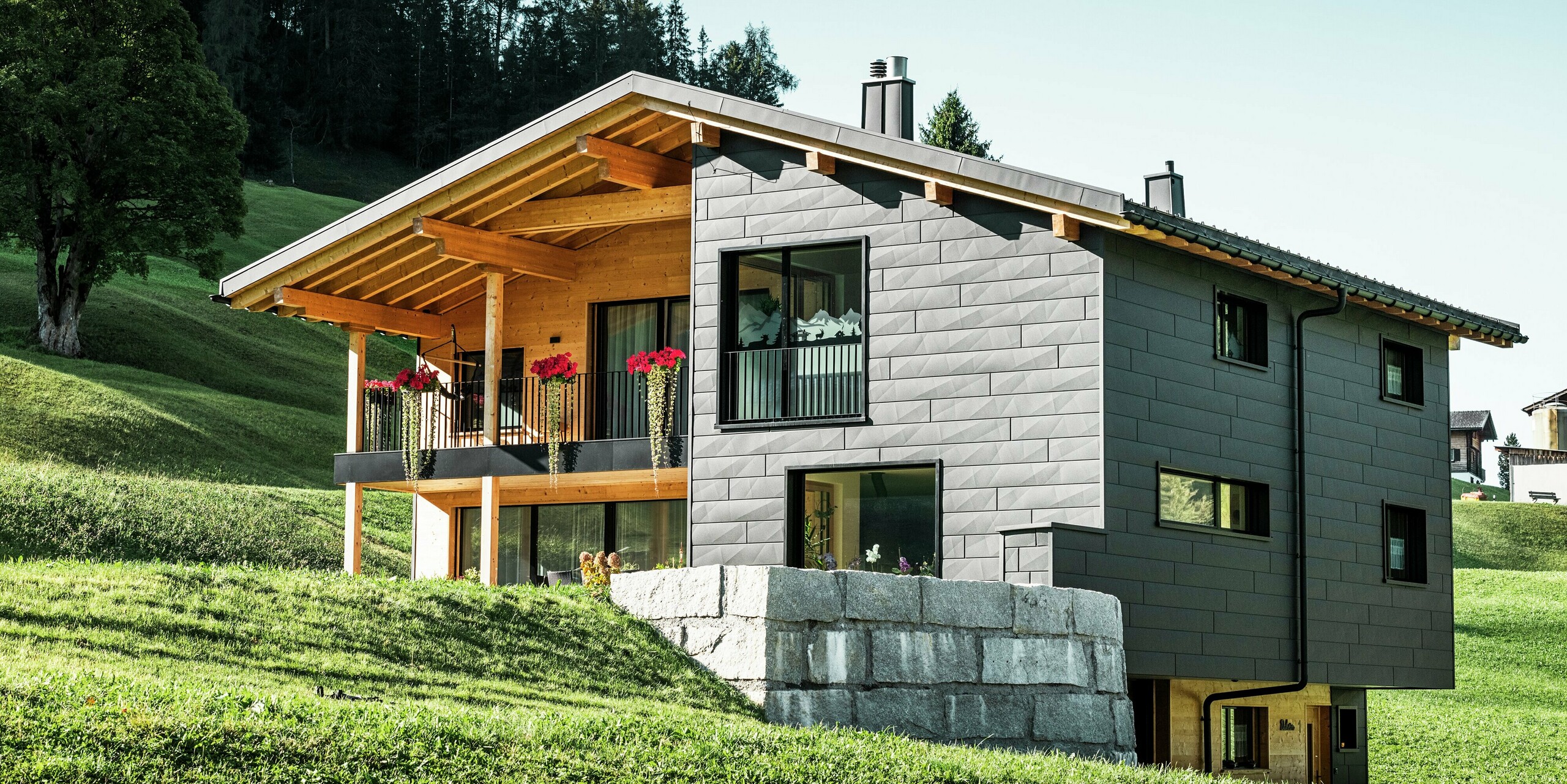 Cette maison individuelle moderne à Ascharina, en Suisse, s'intègre harmonieusement dans le paysage alpin verdoyant. Le bâtiment est entièrement revêtu de panneaux de façade PREFA Siding.X durables et résistants aux intempéries en P.10 noir, qui confèrent à la structure une apparence unique. Le balcon en bois complète parfaitement la coque en aluminium foncé et apporte une touche naturelle.
