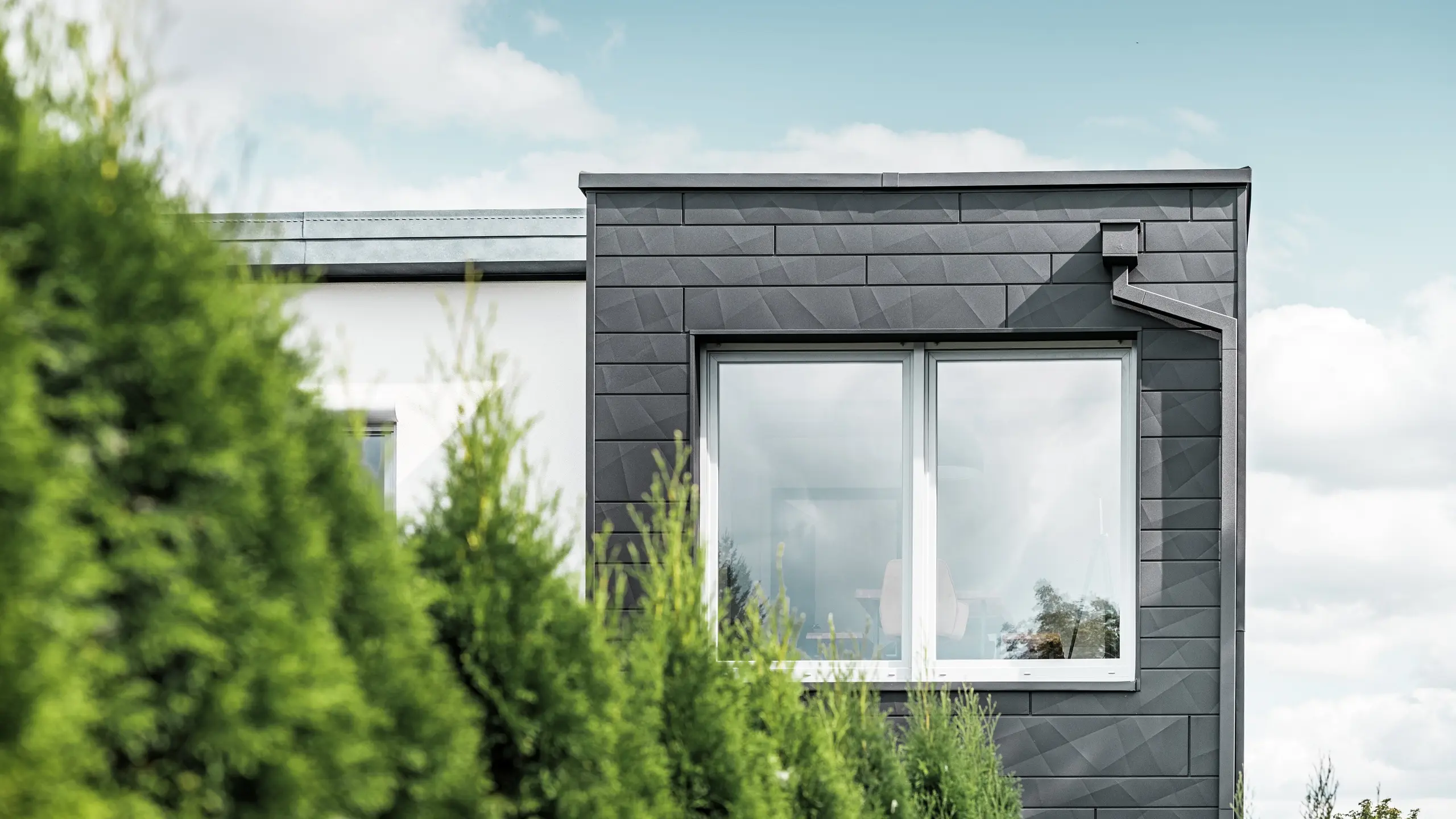 Die klar strukturierten PREFA Siding.X Fassadenpaneele in der Farbe P.10 Schwarz zieren die Außenwand eines Einfamilienhauses in Teuschnitz und verleihen ihm ein einzigartiges Erscheinungsbild. Die PREFA Dachentwässerung, bestehend aus dem Quadratrohr und dem Wasserfangkasten, fügt sich nahtlos in das Design ein. Die grünen Thuja-Bäume im Vordergrund kontrastieren mit der dunklen Blechfassade.