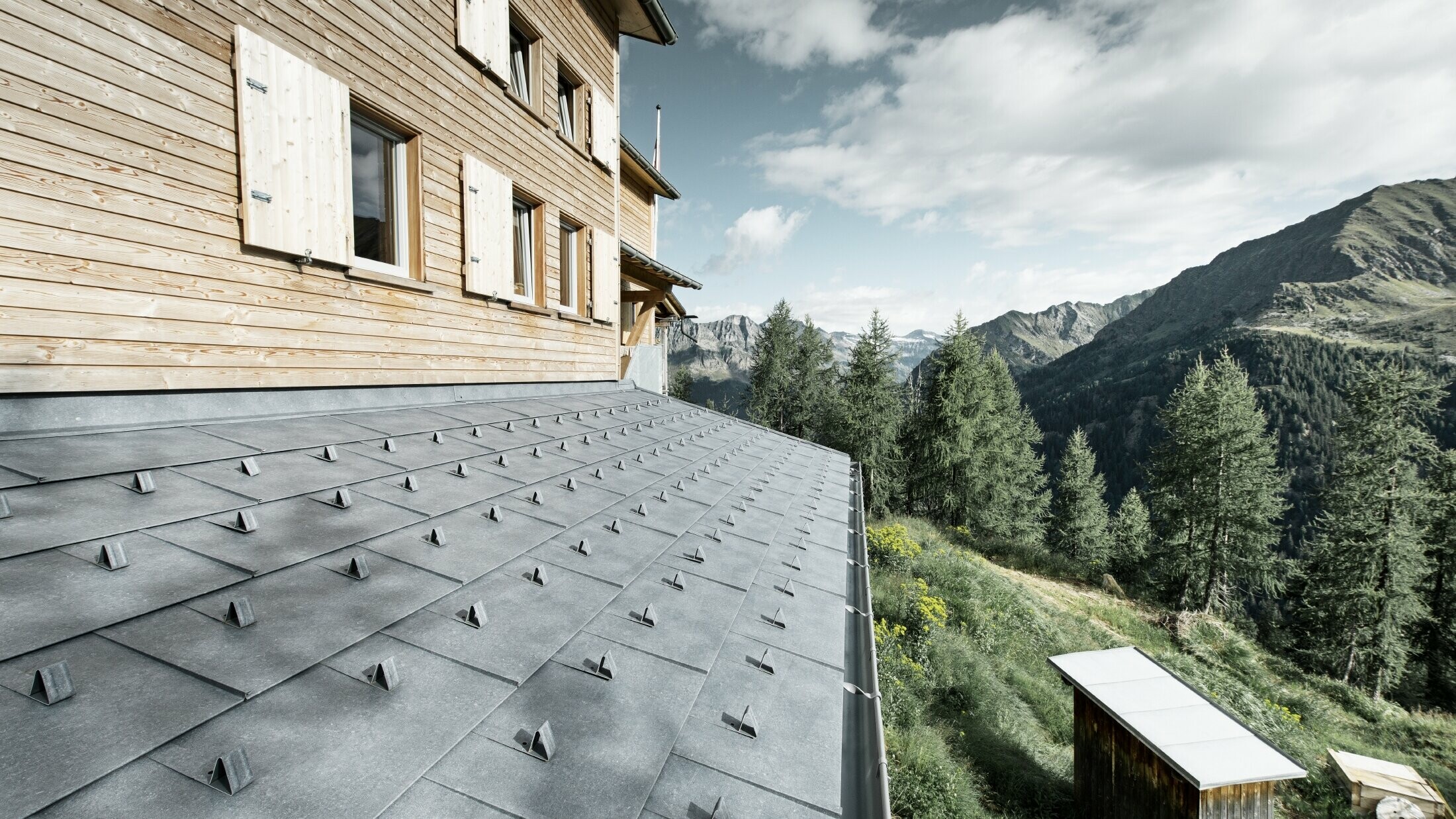 Gros plan sur l’avant-toit d'un refuge en bois. L'avant-toit est recouvert de panneaux d’aluminium FX.12 PREFA couleur gris pierre avec arrêts de neige. La gouttière est maintenue par des brides de renfort.