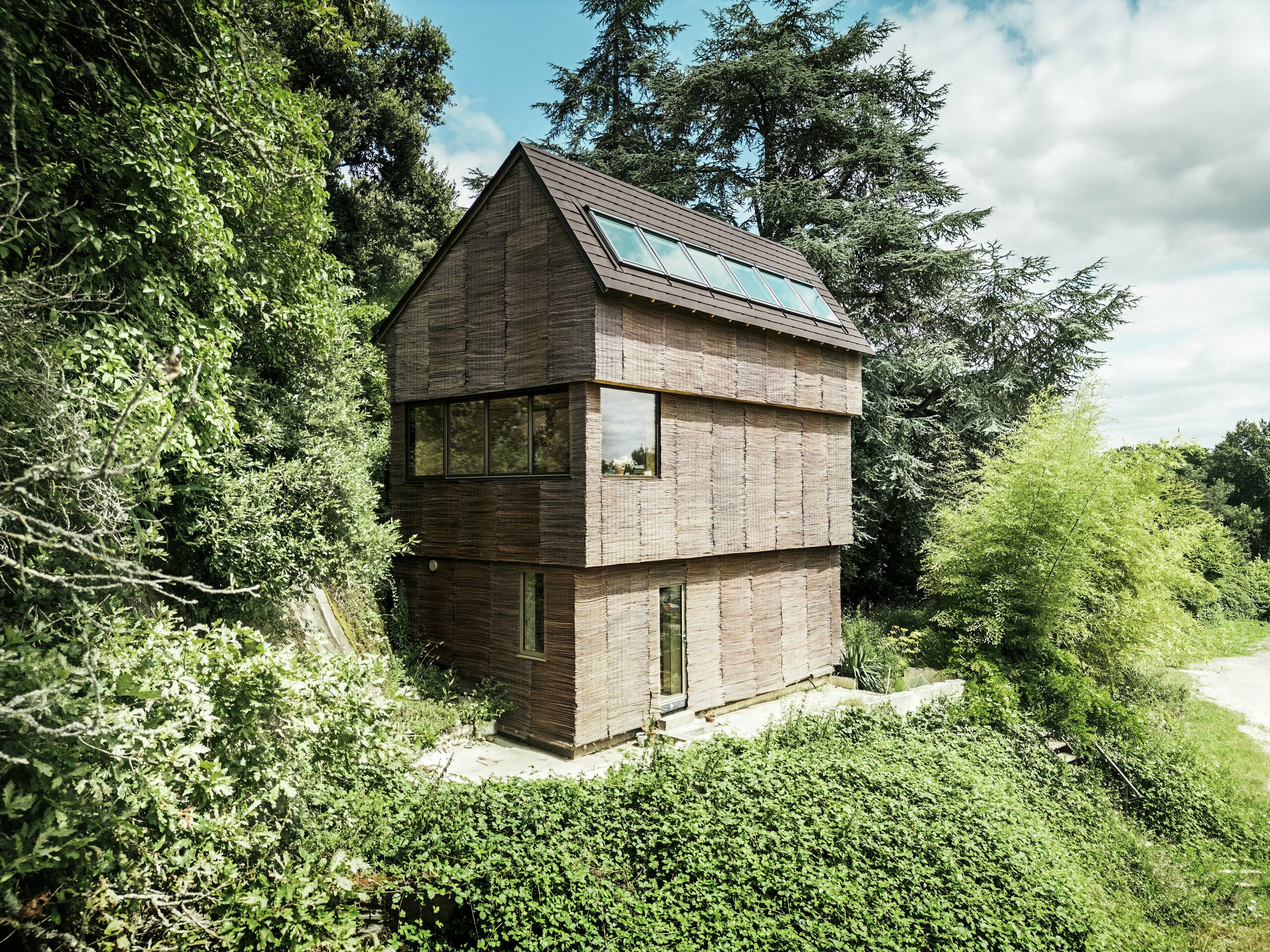 Maison en France avec des bardeaux de toiture PREFA de couleur P.10 brun noisette et une façade en bois de saule, entourée de verdure naturelle – architecture durable et conception respectueuse de l'environnement.