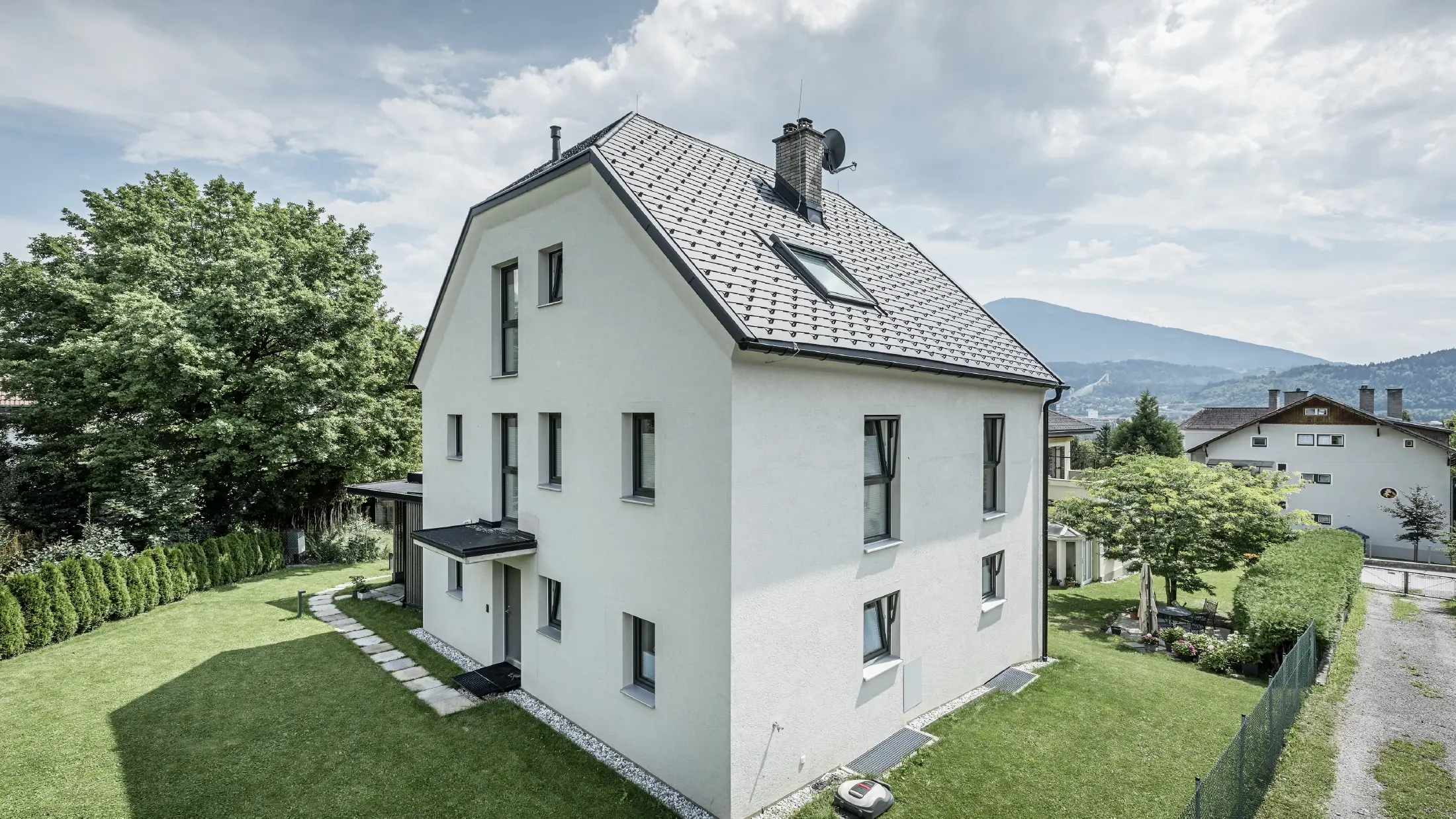 Maison individuelle moderne de trois étages avec une façade claire en crépi et un toit en pente gris sombre en aluminium PREFA. Le bâtiment dispose de plusieurs fenêtres étroites et de gouttières anthracite. Il est situé dans un jardin bien entretenu avec une pelouse, des arbres et des haies. À l'arrière-plan, on aperçoit d'autres maisons et un paysage montagneux.