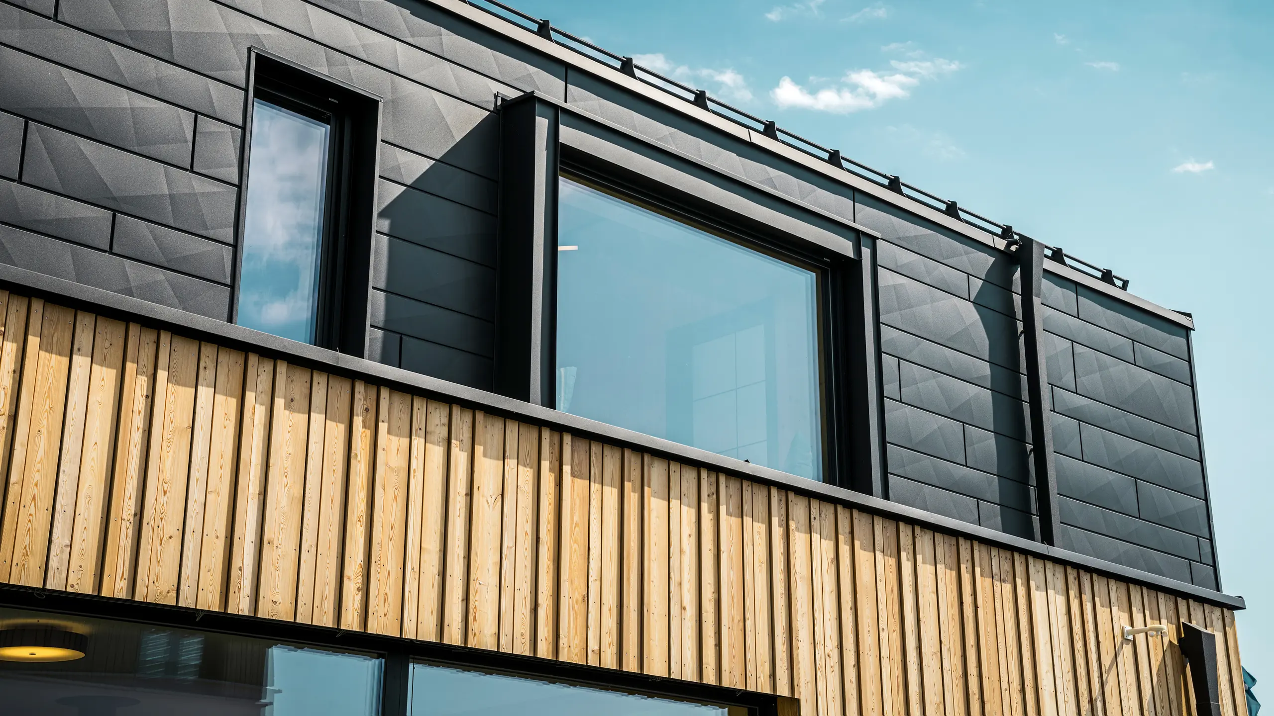 Gros plan sur l'étage supérieur de la maison témoin Autark dans le centre de maisons préfabriquées Blaue Lagune, à Vösendorf (AT). La façade est composée de panneaux en aluminium PREFA Siding.X en P.10 noir qui se distinguent par leur aspect particulier avec différents plis. En dessous se trouve un habillage en bois élégant qui forme un contraste harmonieux avec l'aluminium foncé. De grandes fenêtres laissent entrer beaucoup de lumière à l'intérieur du bâtiment et soulignent le design moderne.