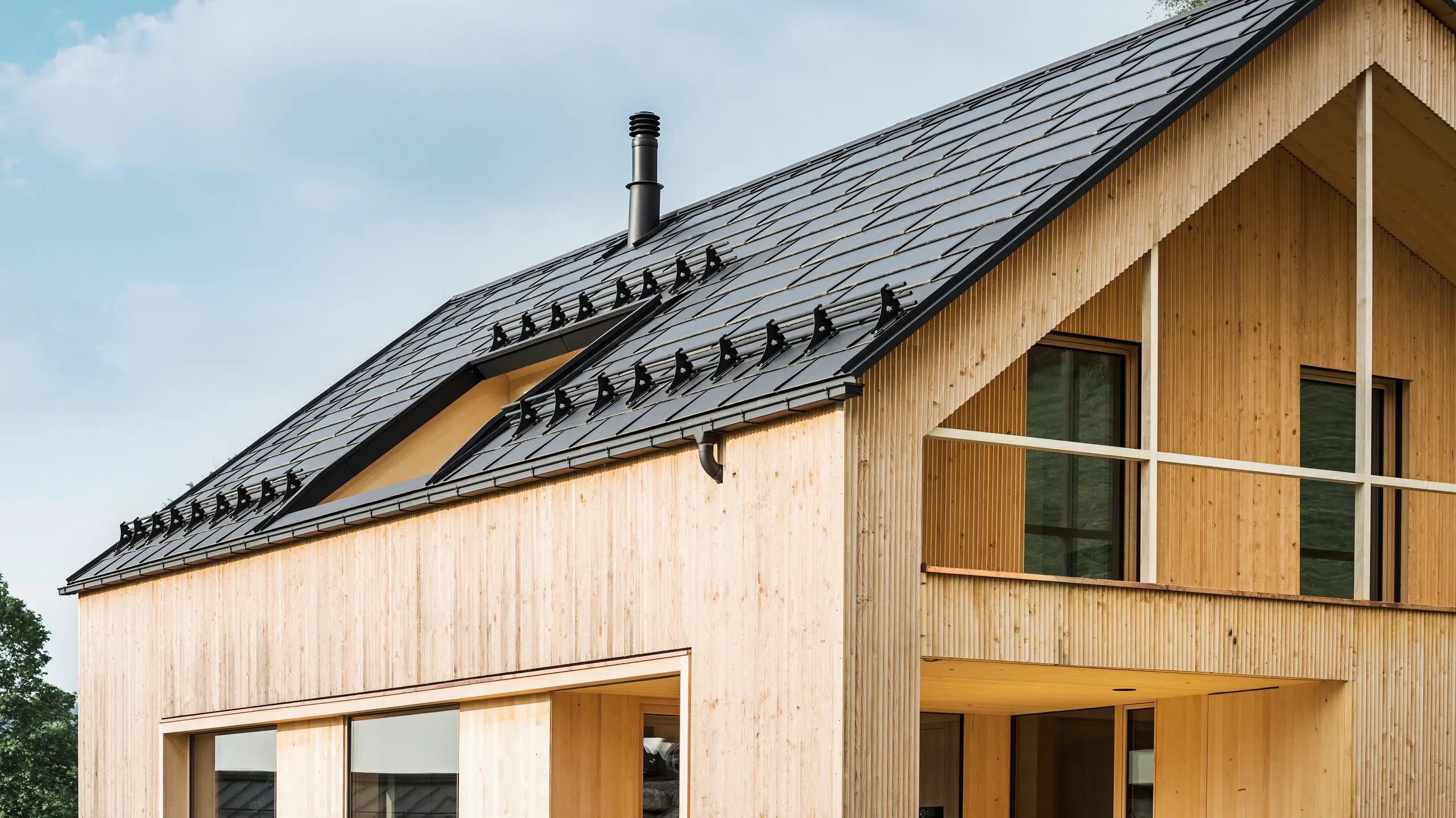 Gros plan sur le pignon d'une maison écologique à Egg, caractérisée par des tuiles solaires PREFA d'un noir élégant, qui s'intègrent harmonieusement au milieu des R.16 PREFA classiques. Les lignes épurées du toit contrastent avec la texture naturelle de la façade en bois, tandis que les tuiles solaires témoignent d'un mode de vie durable. La cheminée subtile s'intègre dans l'ensemble, qui allie un design respectueux de l'environnement et une esthétique rurale. La maison écologique innovante dispose également de deux balcons offrant une vue sur le magnifique paysage montagneux d'Egg dans le Vorarlberg.