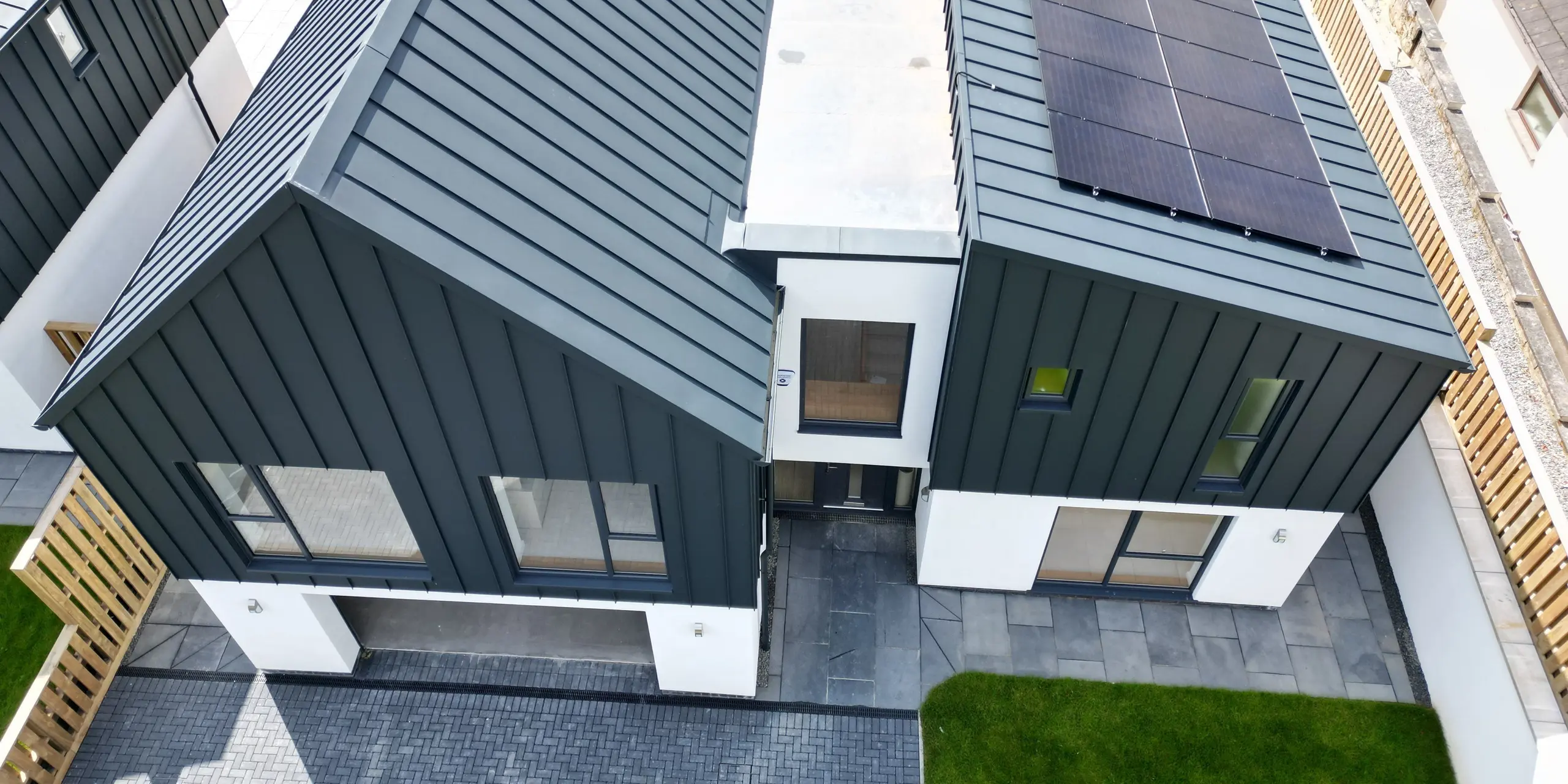 Vue aérienne d'une maison individuelle à Bridgerule, Devon, avec PREFALZ P.10 anthracite comme toiture à joint debout et façade. L'architecture moderne combine l'aluminium durable avec la longévité esthétique, complétée par un système solaire. Un toit à pignon domine l'entrée du garage et la partie droite du bâtiment est dotée d'un toit en appentis. Entre les toits, un pont-bâtiment relie les deux structures.