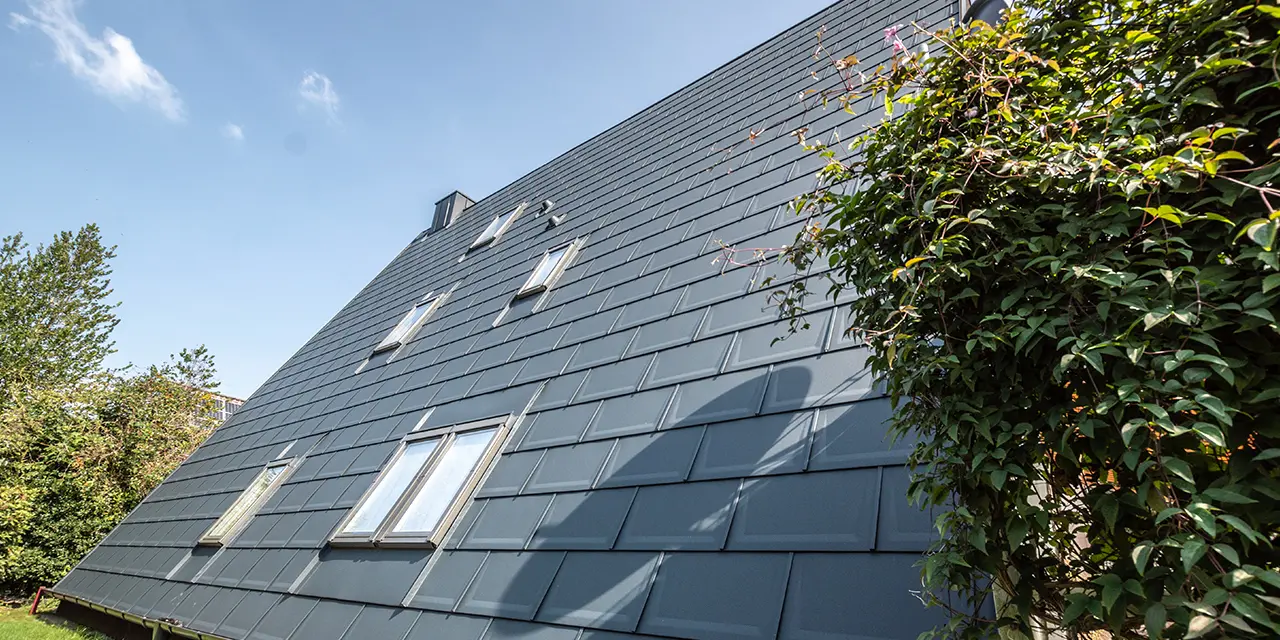 Longue surface de toit d’une maison à ossature en A recouverte de tuiles PREFA couleur P.10 anthracite. Quelques fenêtres de toit sont intégrées dans la surface de toit.