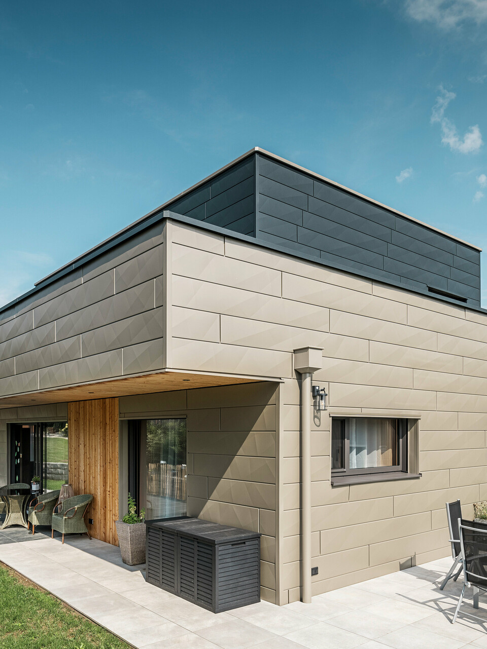 Une maison individuelle moderne à Wippenham, en Autriche, avec une façade en aluminium PREFA bicolore, composée des panneaux de façade Siding.X en bronze et anthracite. La façade présente des lignes et des bords élégants et clairement reconnaissables. Le système d'évacuation des eaux de toiture PREFA harmonieusement intégré avec collecteur d'eau et tuyau d'évacuation est clairement visible et s'intègre parfaitement dans le concept architectural global. Au premier plan, vous pouvez voir une agréable terrasse avec mobilier de jardin, qui souligne le design moderne de la maison nouvellement construite.